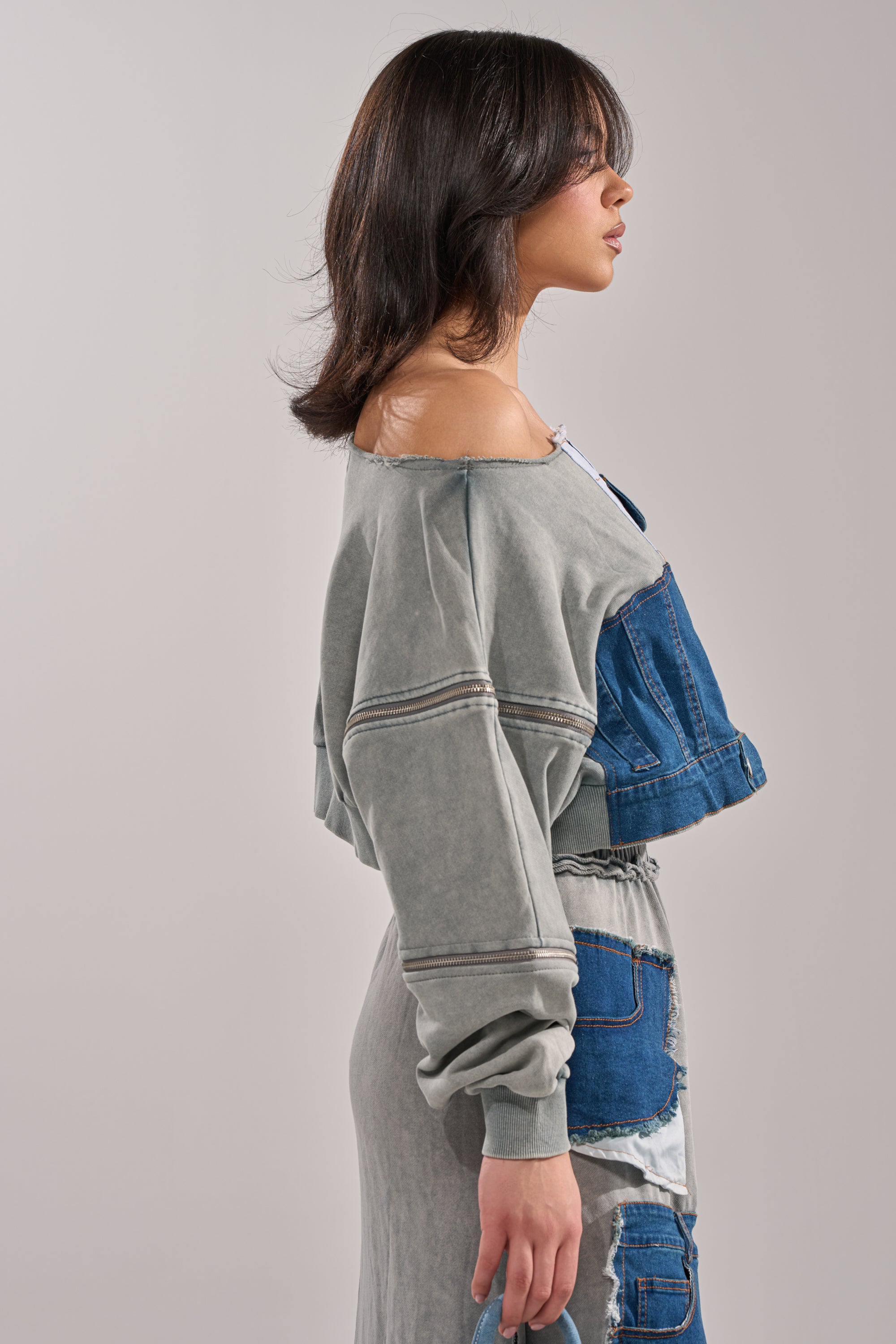 A woman with shoulder-length brown hair stands in profile against a plain light background, wearing the DENIM DREAMS ZIPPER DETAIL SWEATSHIRT in gray denim patchwork with matching skirt and denim pockets.