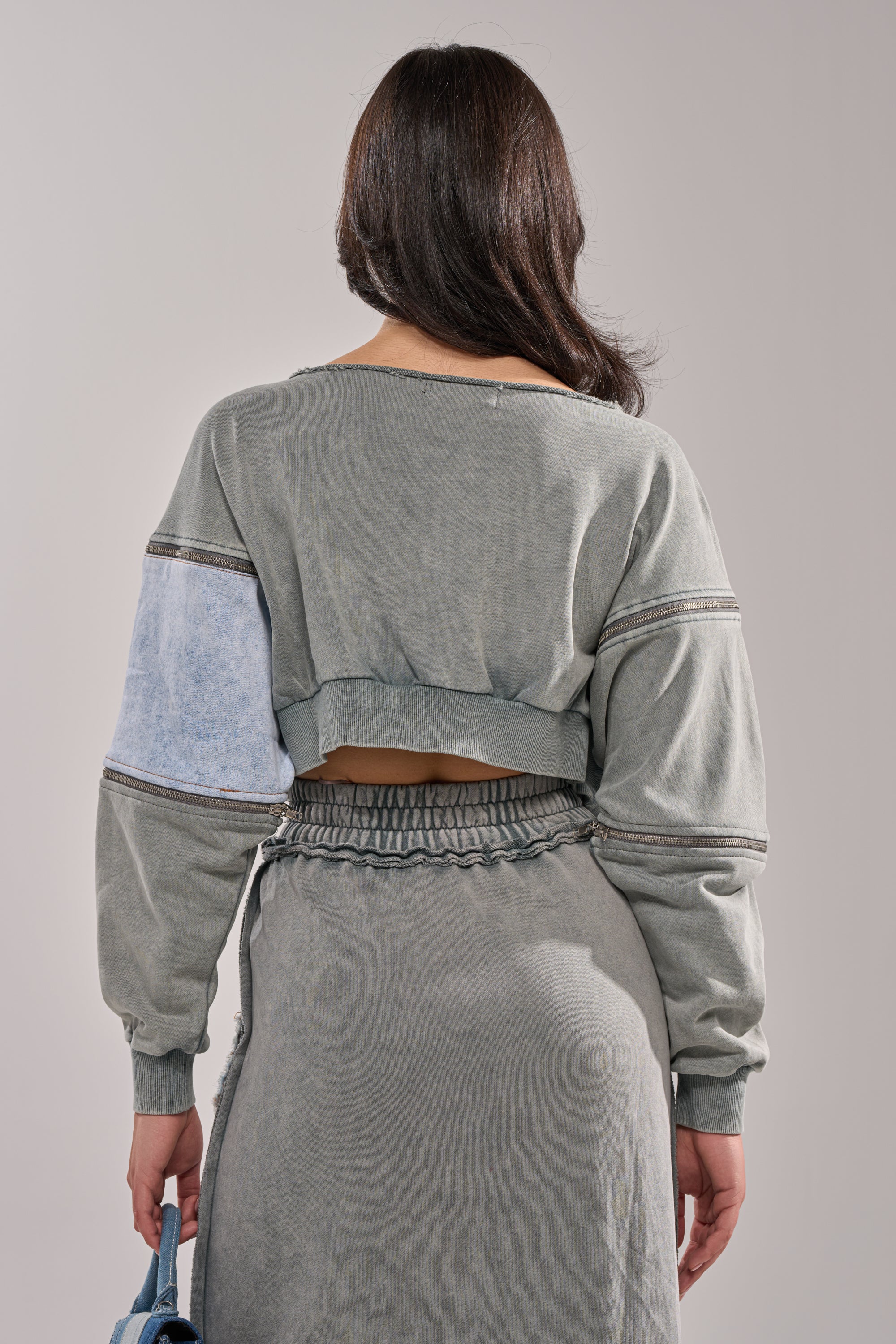 A woman stands facing away, wearing the DENIM DREAMS ZIPPER DETAIL SWEATSHIRT, a gray cropped denim patchwork top with a light blue patch on one sleeve, paired with a matching high-waisted skirt and holding a light blue handbag in her left hand.