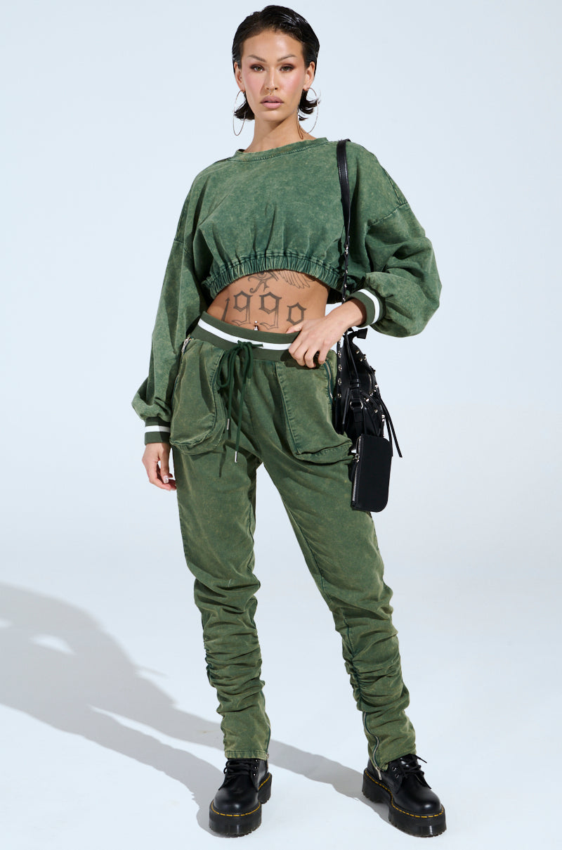 On a neutral background, someone wears the DUSTIN GREEN JOGGER, characterized by a cropped sweatshirt and high-rise joggers. Tattoos peek above the waistband while they carry a black bag and wear black boots, with the look completed by a mineral wash touch.