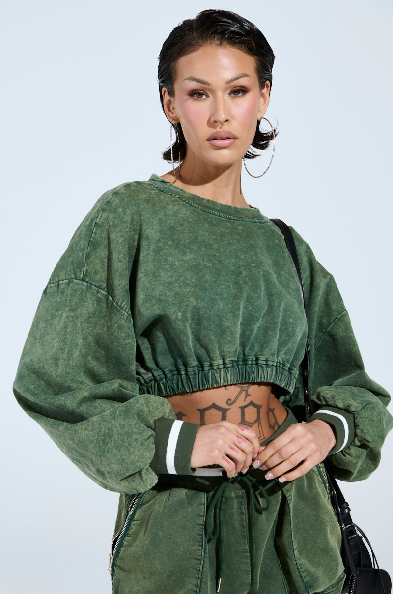 The person is styling the green DUSTIN CROPPED CREW sweatshirt and matching pants, with slicked-back hair, hoop earrings, bold eyeliner, holding a black bag. A tattoo adorns their midriff against a plain white background.