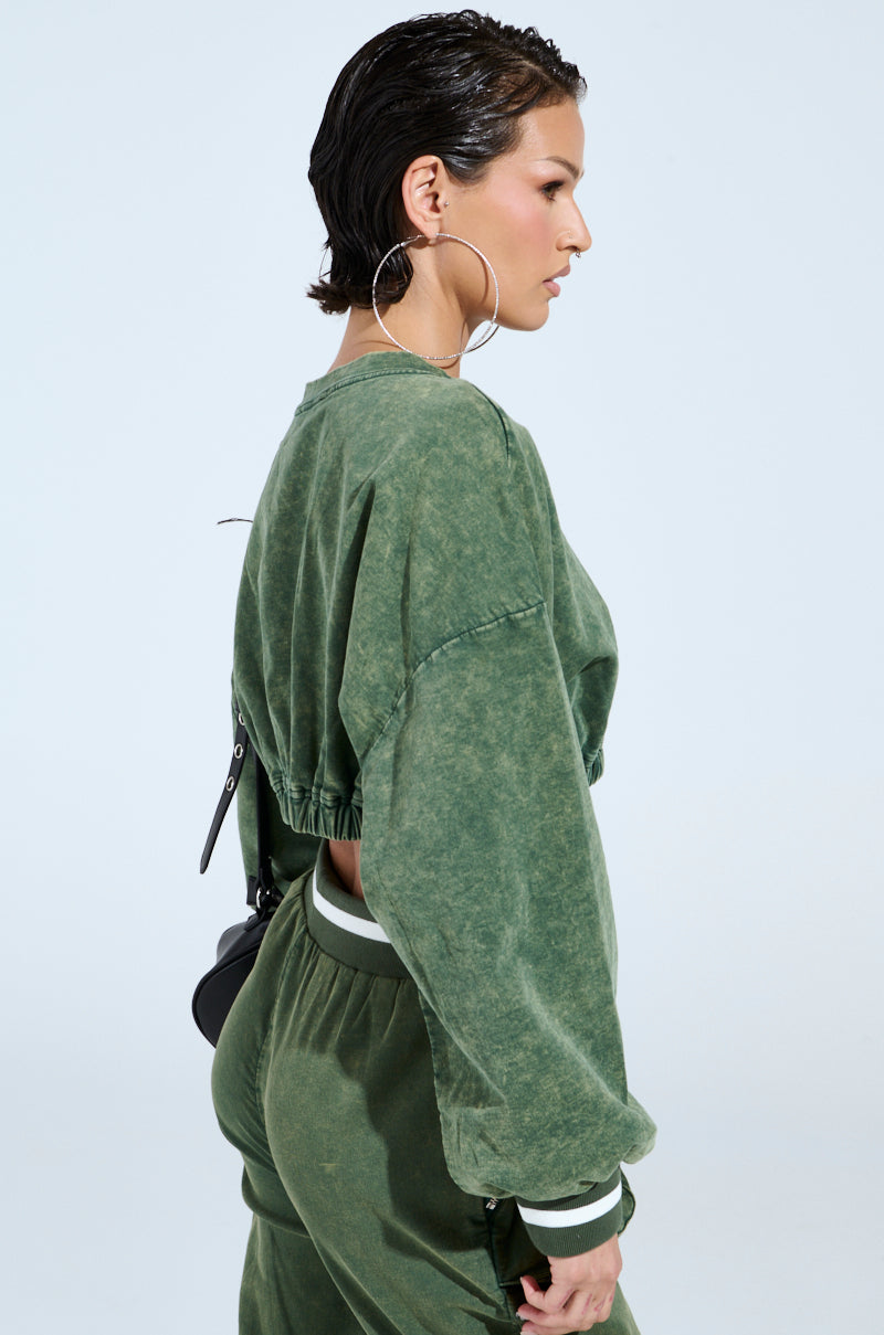 Someone with short hair is in a green DUSTIN CROPPED CREW and matching pants. They're wearing a black strap across their back and large hoop earrings. The light gray background accentuates the outfit's oversized silhouette.