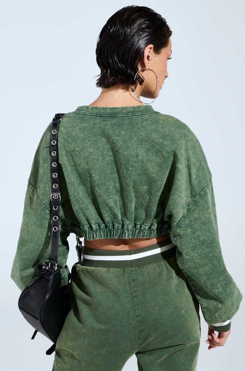A person with short dark hair wears the DUSTIN CROPPED CREW sweatshirt and matching green pants, embracing an oversized silhouette. They carry a black shoulder bag, sport large hoop earrings, and stand against a soft light blue background.