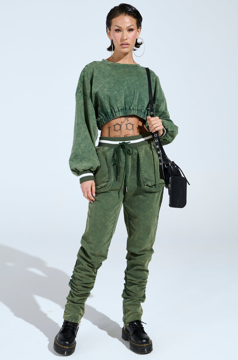A person wears the DUSTIN CROPPED CREW, a green mineral wash cropped sweatshirt with matching joggers. They accessorize with black boots, hoop earrings, and a black shoulder bag, revealing their "1999" tattoo on their midriff.