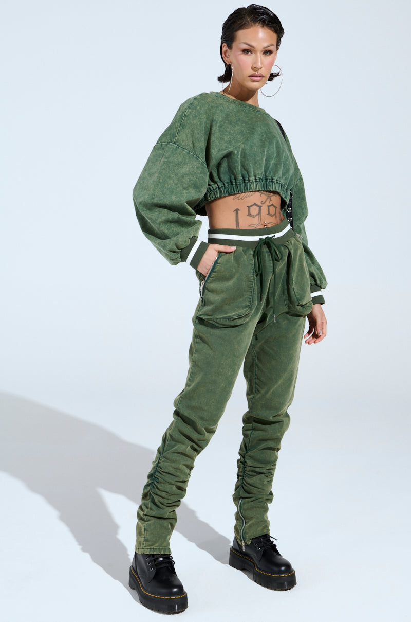 Confidently wearing a DUSTIN CROPPED CREW green two-piece with an oversized silhouette, they sport a long-sleeve crop top and matching pants, complemented by black boots and large hoop earrings. A tattoo adorns their torso against a plain light background.