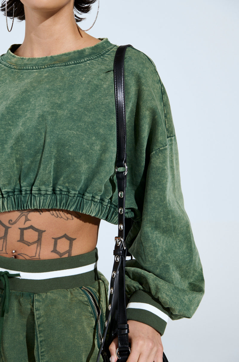 Person showcases a DUSTIN CROPPED CREW in green, featuring an oversized silhouette with long sleeves and matching pants. A torso tattoo appears beneath, accessorized with large hoop earrings and a black strap over the shoulder, set against a mineral wash backdrop in plain light blue.
