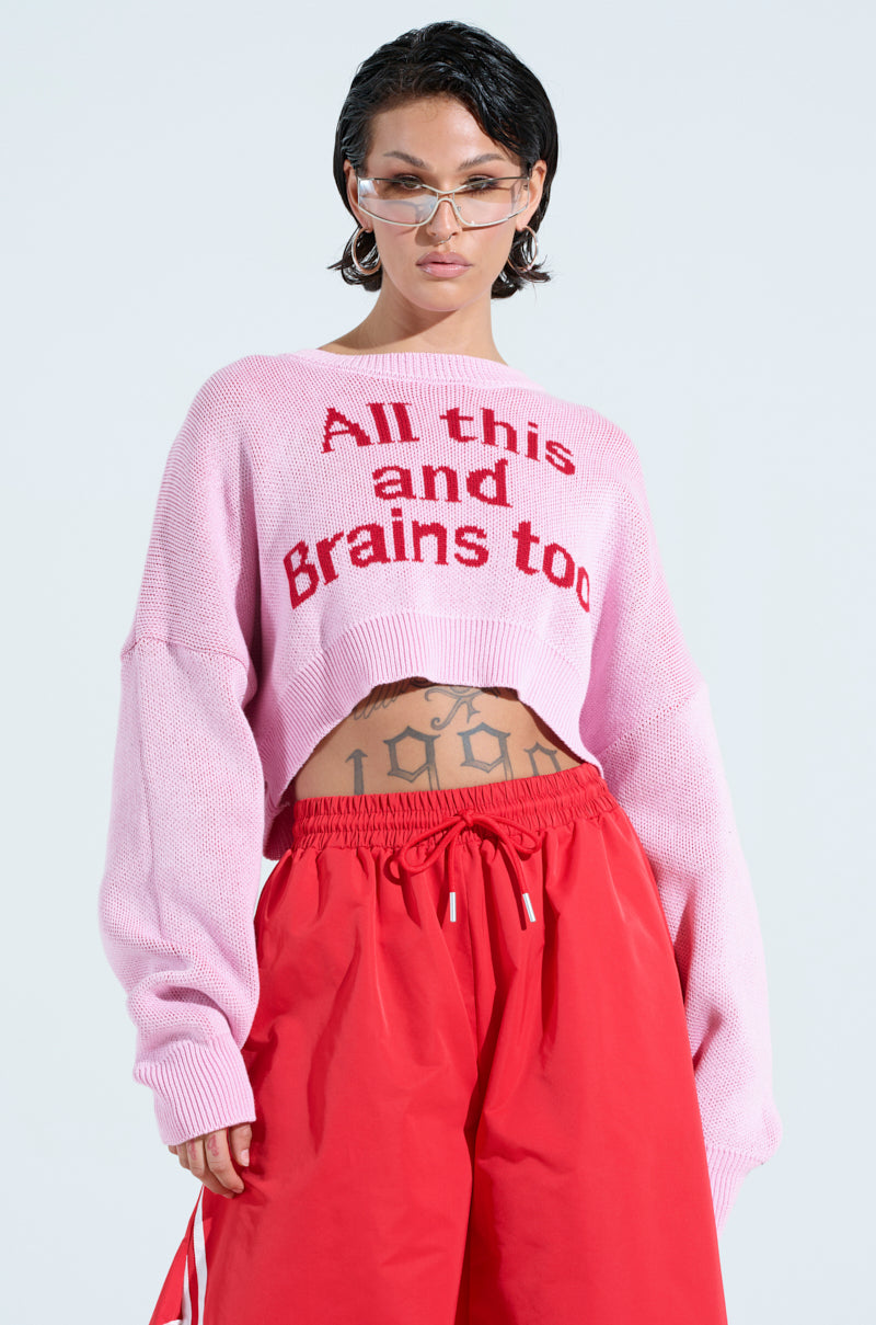 Wearing the ALL THIS AND BRAINS TOO CROPPED SWEATER in pink with red pants, their short hair, stylish sunglasses, and visible tattoos create a striking look against the plain white background.