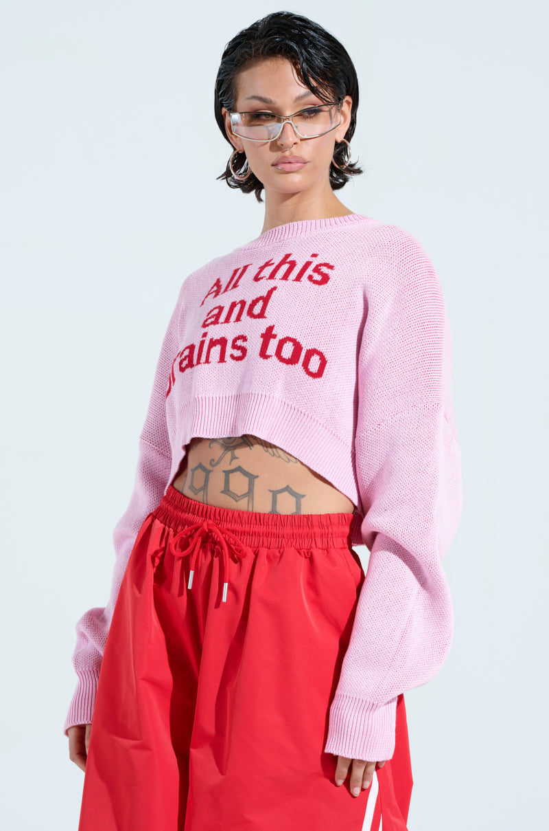 A person with short hair wears the ALL THIS AND BRAINS TOO CROPPED SWEATER in light pink, paired with red high-waisted pants and transparent glasses. Tattoos adorn their abdomen as they stand against a plain background.