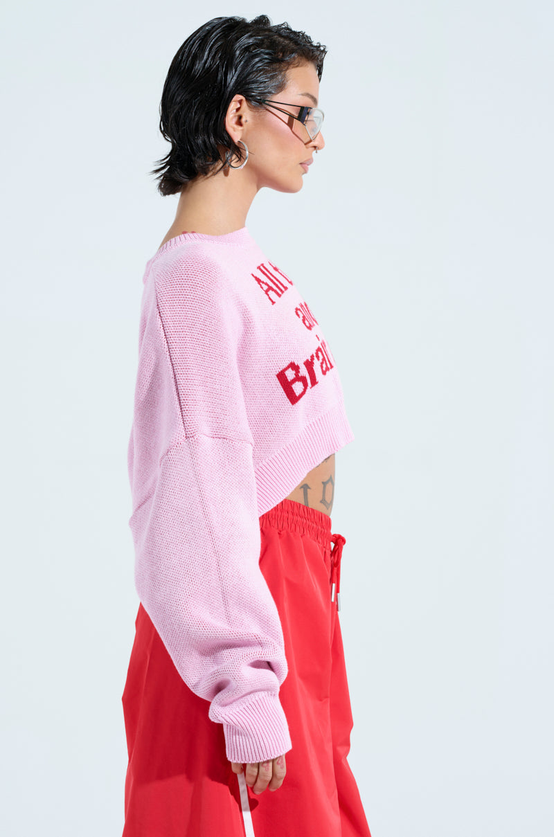 A person with short dark hair is wearing glasses and the ALL THIS AND BRAINS TOO CROPPED SWEATER in light pink with red text, paired with red pants. They stand sideways against a plain light background.