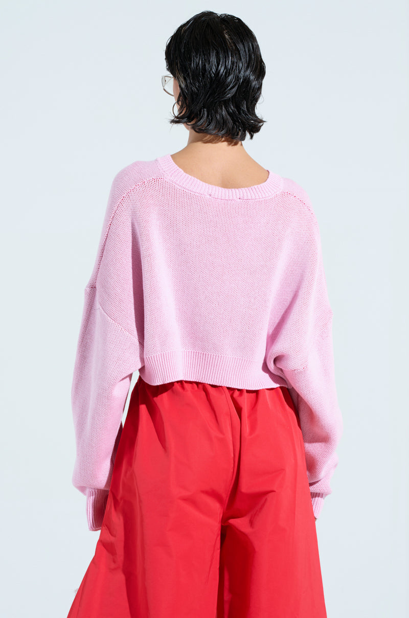 A person with short black hair is wearing an ALL THIS AND BRAINS TOO CROPPED SWEATER in light pink and red wide-legged pants, standing with their back to the camera against a light blue background.