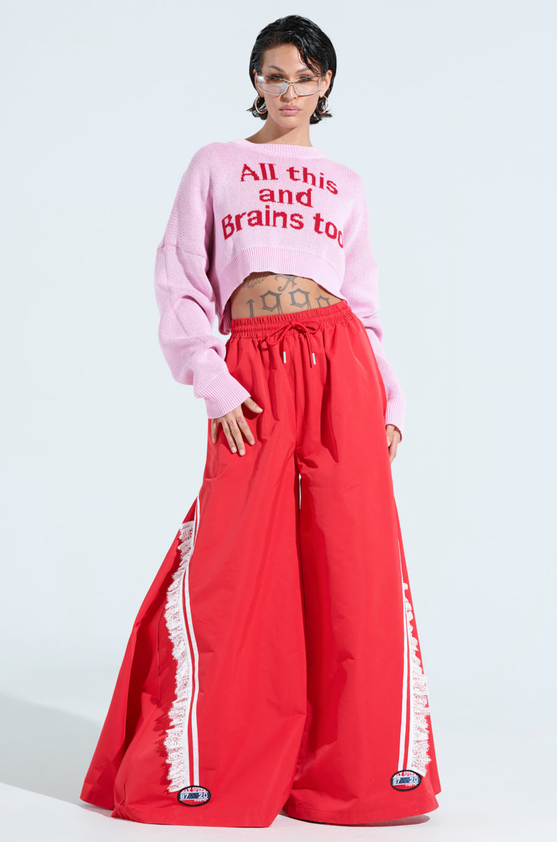 A person confidently poses in an ALL THIS AND BRAINS TOO CROPPED SWEATER, bright red wide-leg pants with white stripe details, set against a plain light background.