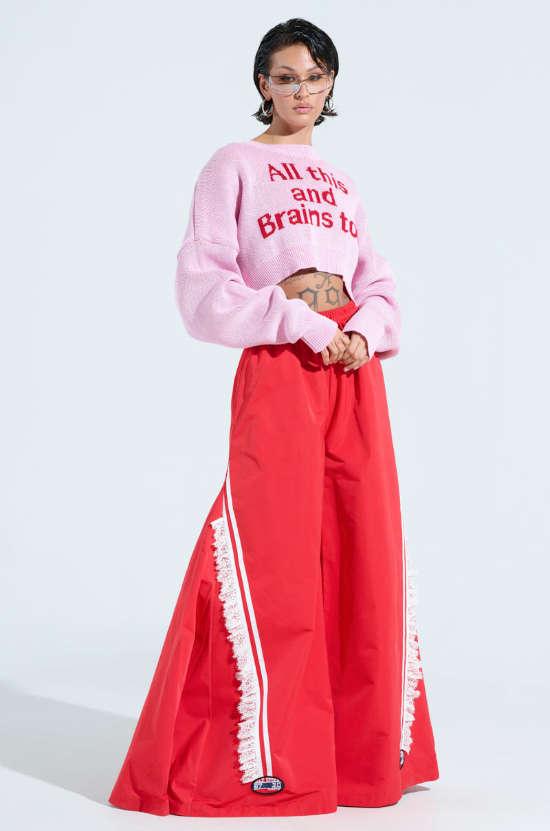 A person with tattoos on their stomach poses against a light background, wearing the ALL THIS AND BRAINS TOO CROPPED SWEATER, an oversized pink graphic sweater, red wide-legged pants with white lace trim, and sunglasses.