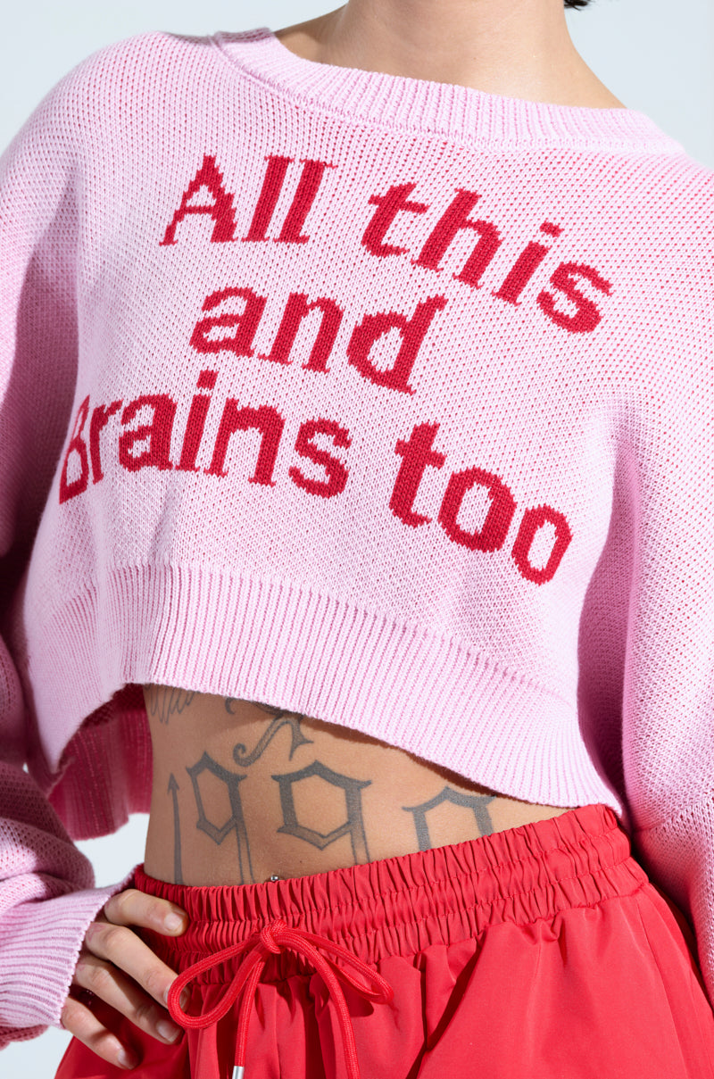Wearing the "ALL THIS AND BRAINS TOO" cropped sweater, styled with red pants featuring a loose drawstring, adds flair against a light gray background. Visible tattoos, including numbers on their stomach, contribute to an edgy vibe.
