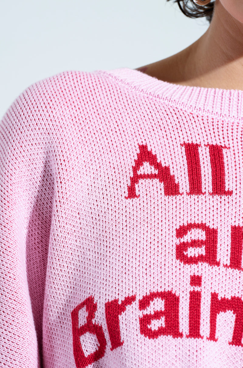 Close-up of a person wearing the "ALL THIS AND BRAINS TOO CROPPED SWEATER," featuring bold red text partially visible on a pink graphic fabric. The textured material and oversized silhouette create a striking contrast between the light pink and vibrant red lettering.