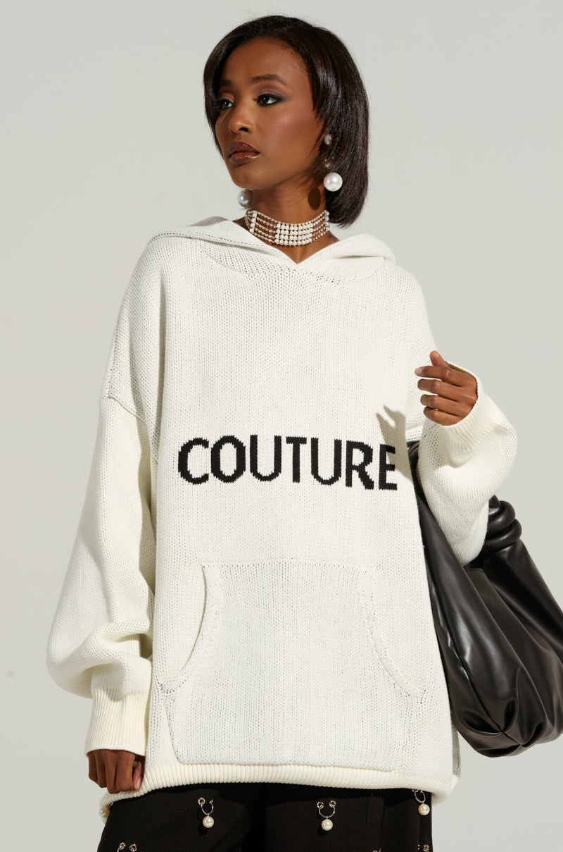 A person is modeling a COUTURE OVERSIZED HOODED SWEATER in ivory, featuring the word "COUTURE" printed on the front. They are accessorized with a pearl choker necklace and pearl earrings, carrying a large black bag and wearing black pants adorned with metallic embellishments.