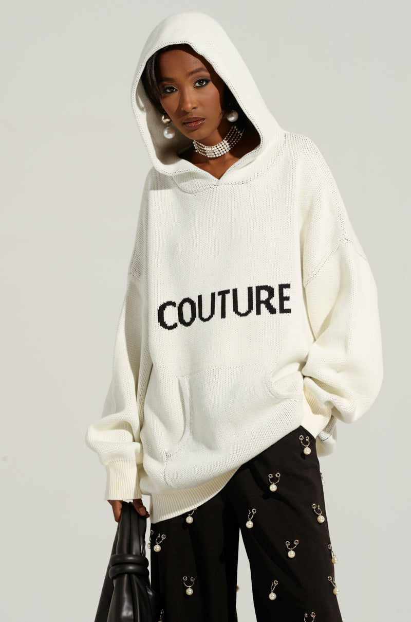 A woman wearing a Couture Oversized Hooded Sweater in ivory, paired with black pants adorned with small decorative elements. She also has a choker necklace, large pearl earrings, and is holding a black handbag. The background is plain light gray.