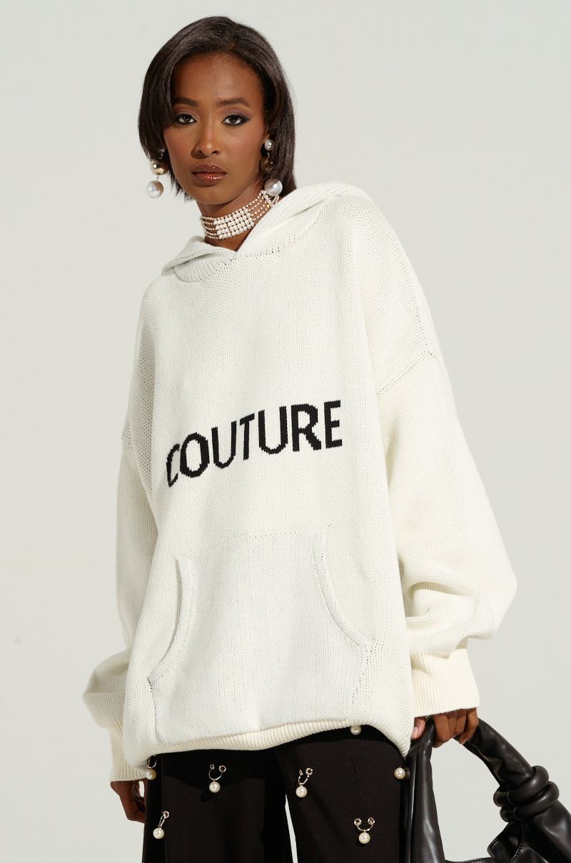 A woman with a short bob haircut poses confidently in an ivory COUTURE OVERSIZED HOODED SWEATER that reads "COUTURE" in bold black letters. She accessorizes with a pearl necklace and pearl earrings. Holding a textured black bag, she completes the look with black pants featuring decorative embellishments.