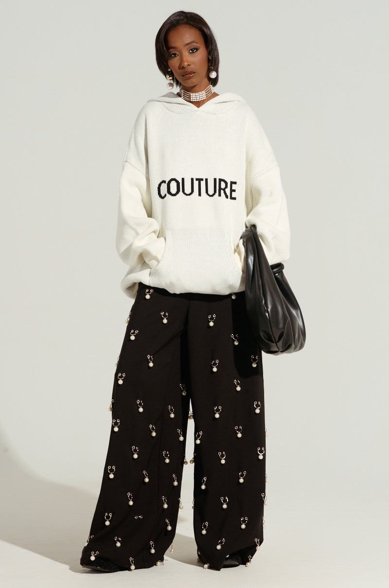 A woman is dressed in a striking ivory COUTURE OVERSIZED HOODED SWEATER emblazoned with the word "COUTURE." She pairs it with cozy double-knit, wide-leg black pants adorned with small decorative patterns. Holding a large black handbag and wearing a choker necklace, she exudes effortless style.