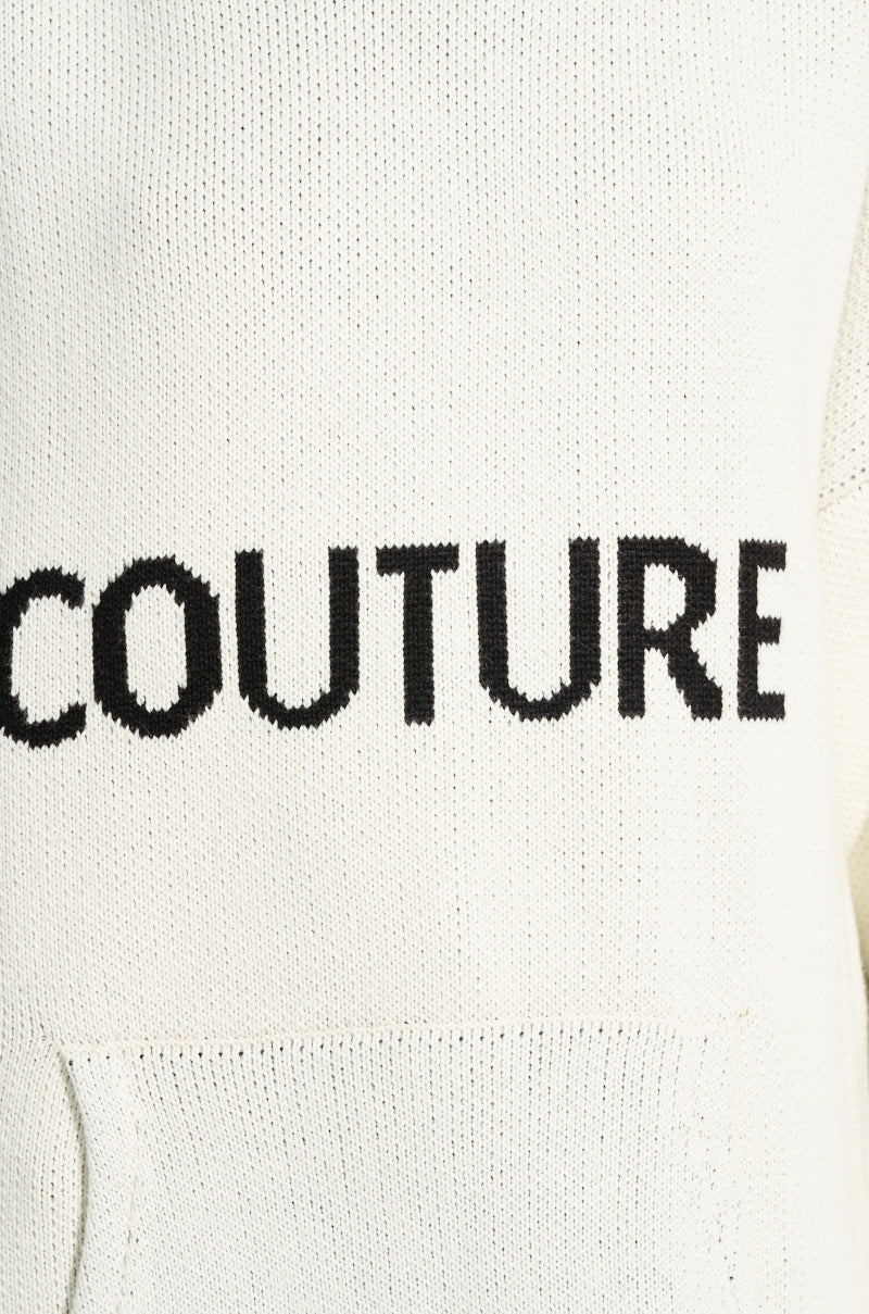 Close-up of a white knitted fabric with the word "COUTURE" stitched in black letters across the center. The fabric, part of the COUTURE OVERSIZED HOODED SWEATER, features a slight ribbed texture and cozy doubled knit, with a pocket hem visible at the bottom.