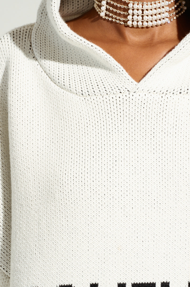 A close-up of a person wearing the ivory COUTURE OVERSIZED HOODED SWEATER, featuring a cozy double-knit texture and prominent weave details. The individual's lower face and neck are visible, adorned with a multi-strand pearl choker.