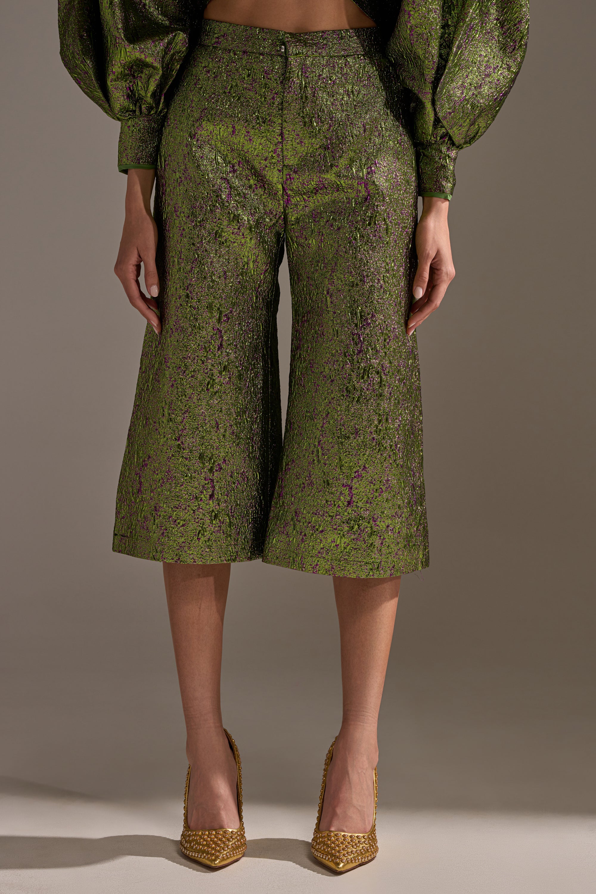 A person stands against a plain background in LAYLA BROCADE CULOTTES—green metallic culottes with a subtle purple pattern—paired with a matching long-sleeved top and pointed gold heels, making them the perfect statement for holiday parties.