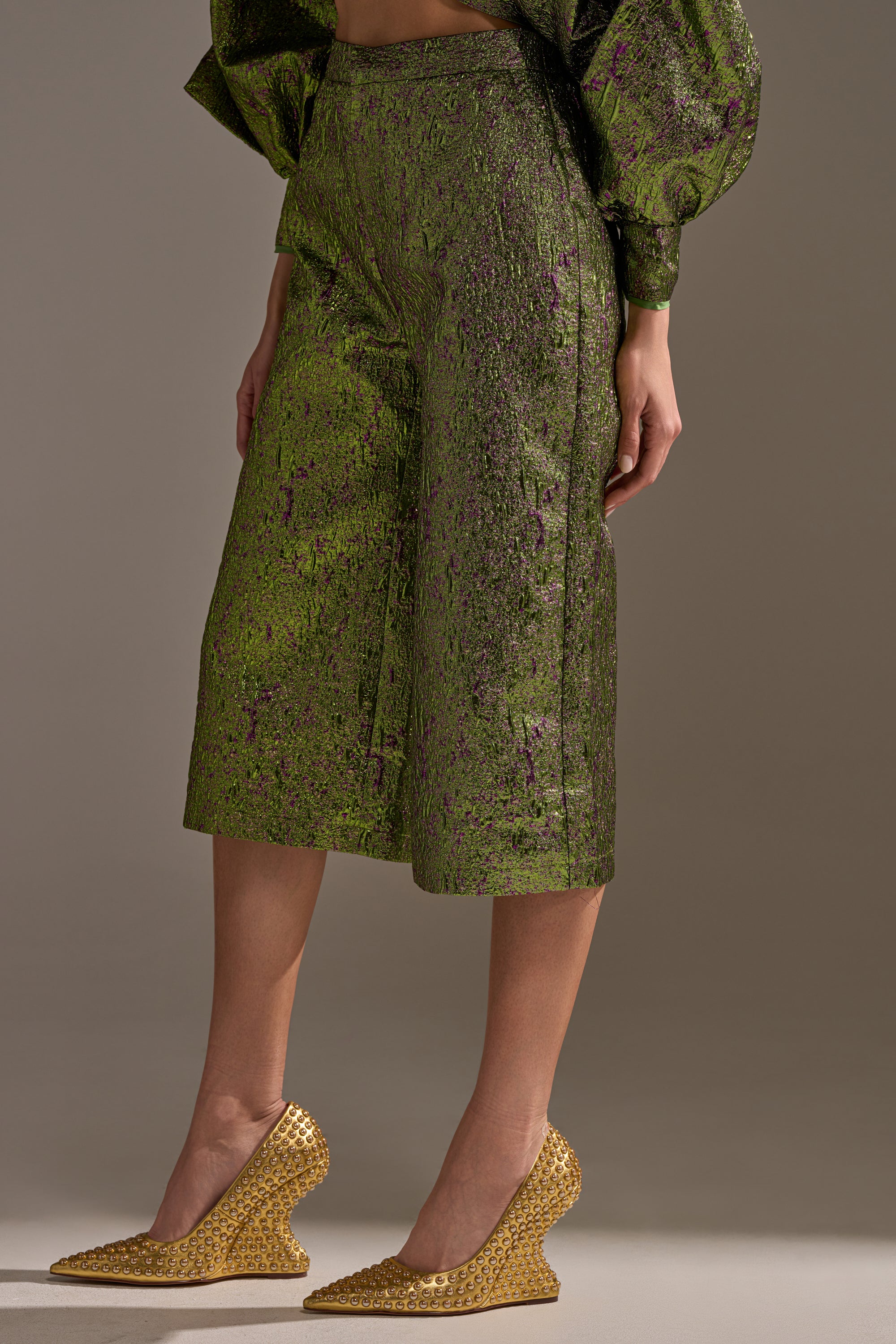 A person wears the LAYLA BROCADE CULOTTES—shin-length, green metallic with a textured pattern—paired with a matching top and gold woven wedge shoes, standing against a neutral background. Ideal for making an entrance at any holiday party.