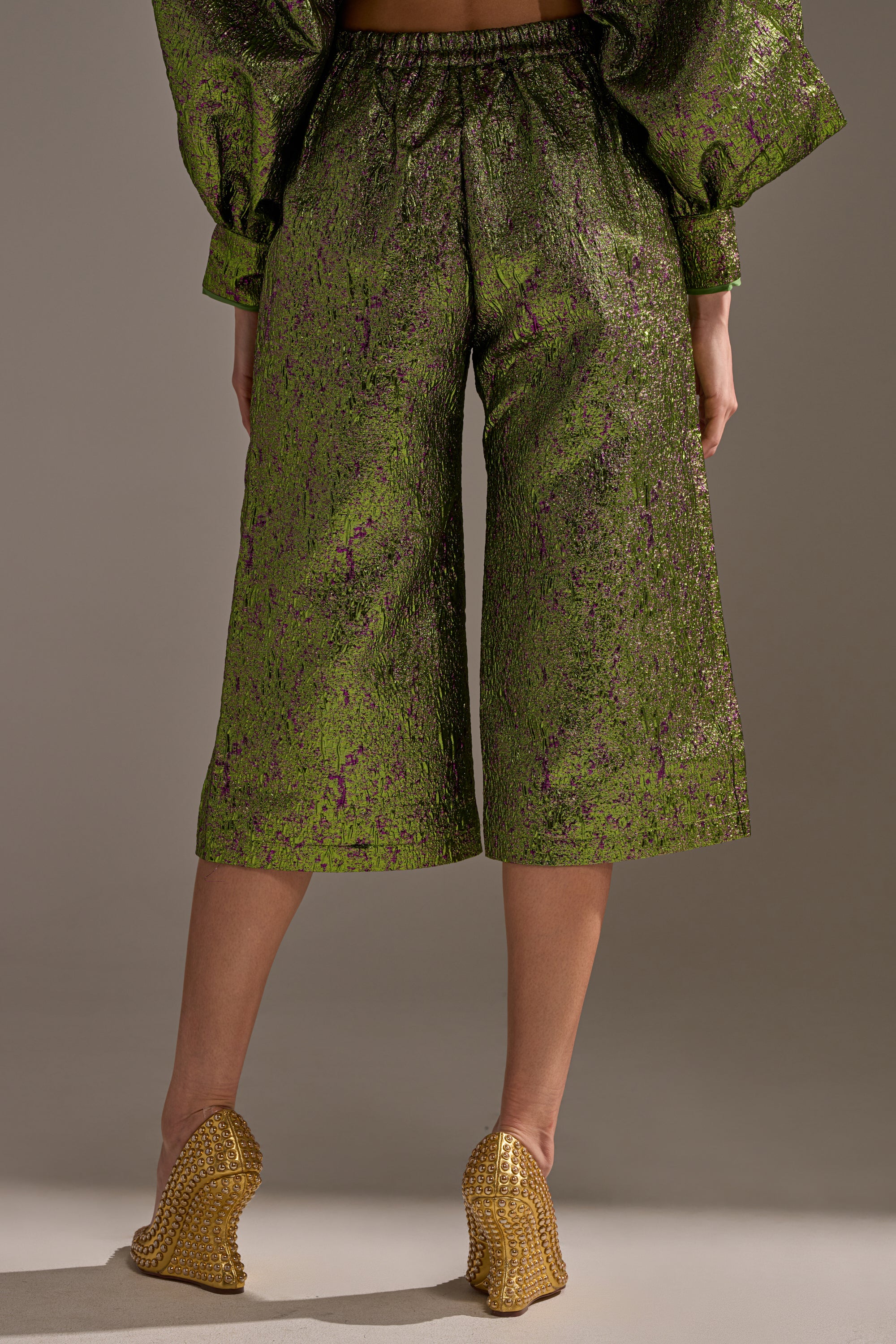 Someone stands against a plain background in the LAYLA BROCADE CULOTTES—shiny green metallic brocade pants with a purple pattern—paired with gold high heels, creating a perfect holiday party outfit.