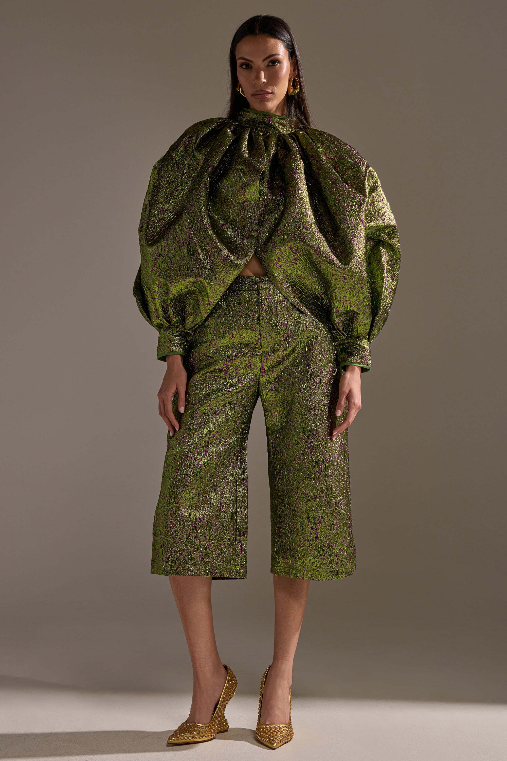 A woman faces forward in the LAYLA BROCADE CULOTTES—shiny green and gold metallic brocade pants—styled with voluminous sleeves, gold pointed-toe heels, and bold earrings for a striking holiday party look.