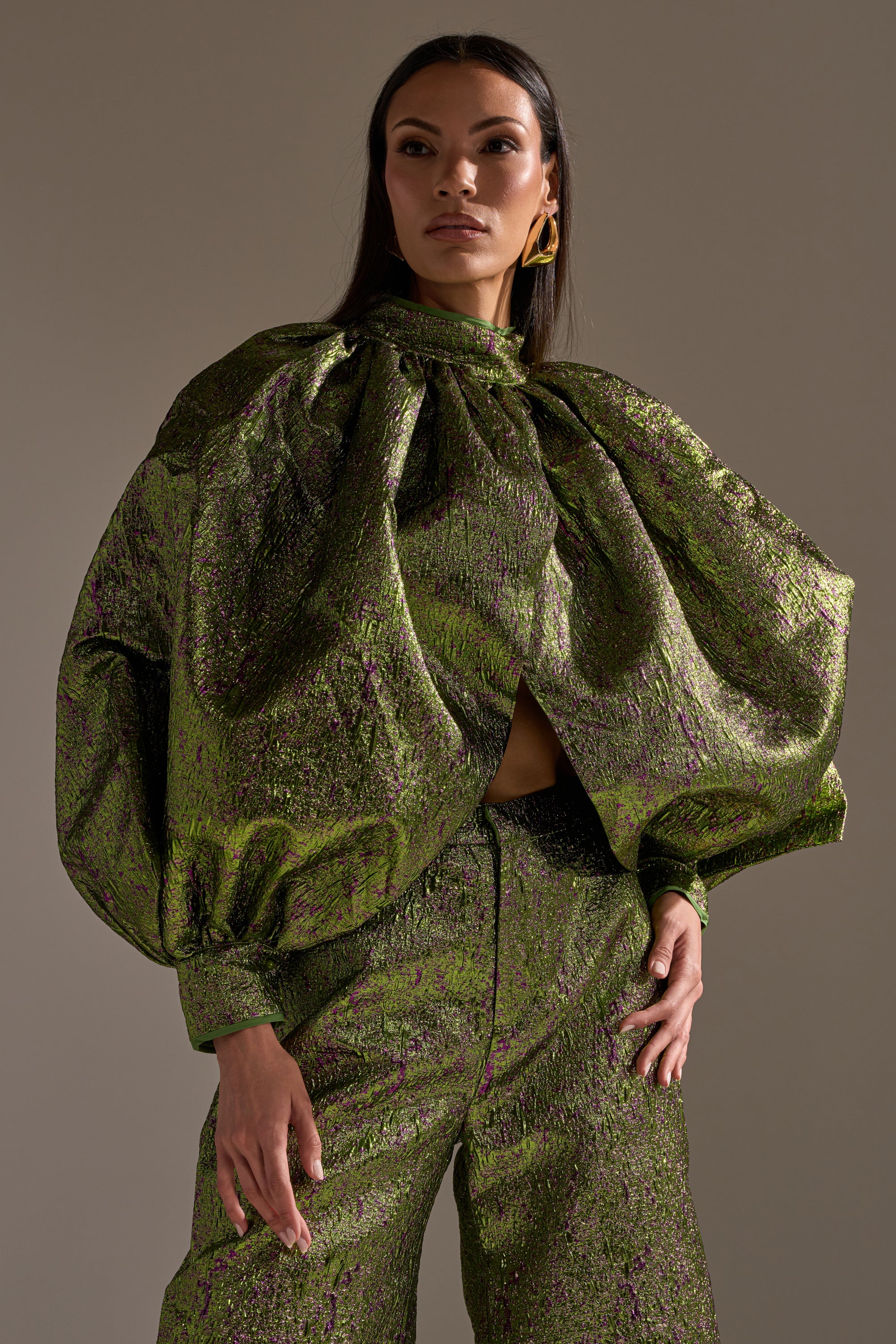 A woman poses confidently in the LAYLA BROCADE PUFF SLEEVE MOCK NECK TOP, featuring metallic fabric and dramatic sleeves, paired with high-waisted green and purple pants against a plain backdrop.
