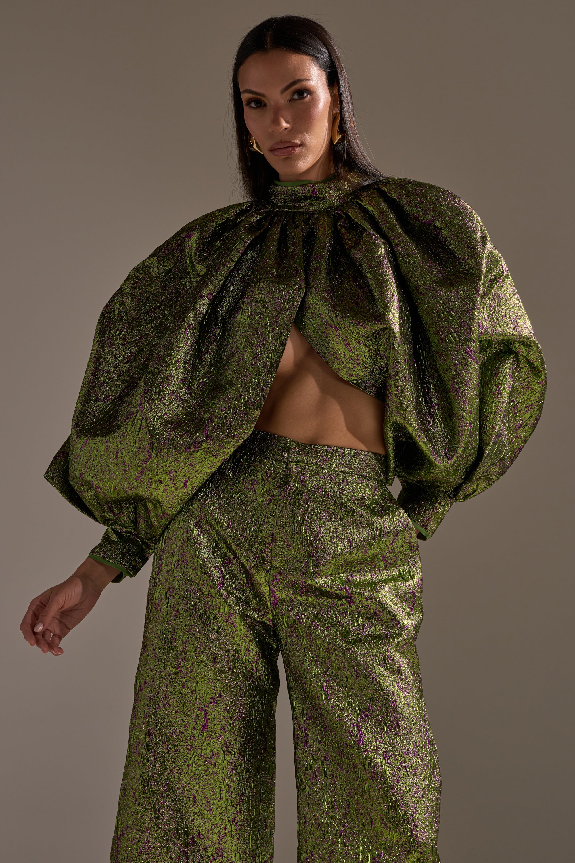 A woman poses in the LAYLA BROCADE PUFF SLEEVE MOCK NECK TOP, featuring a metallic finish, puff sleeves, mock neck, and cropped silhouette that reveals her midriff, styled with high-waisted wide-leg pants.