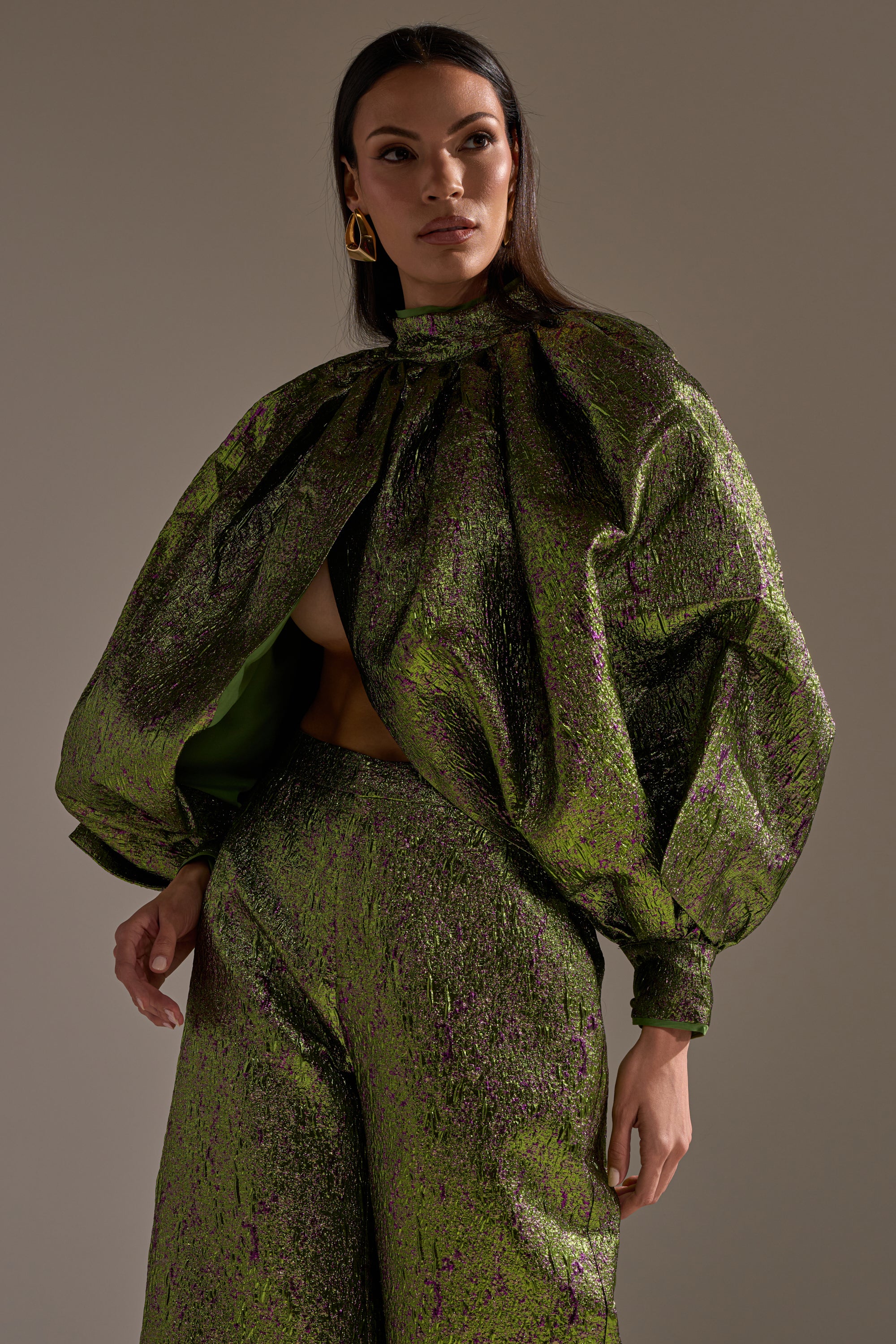 She wears the LAYLA BROCADE PUFF SLEEVE MOCK NECK TOP with voluminous sleeves, paired with wide-leg pants and bold gold earrings, posing confidently against a neutral background to highlight her striking cropped silhouette.