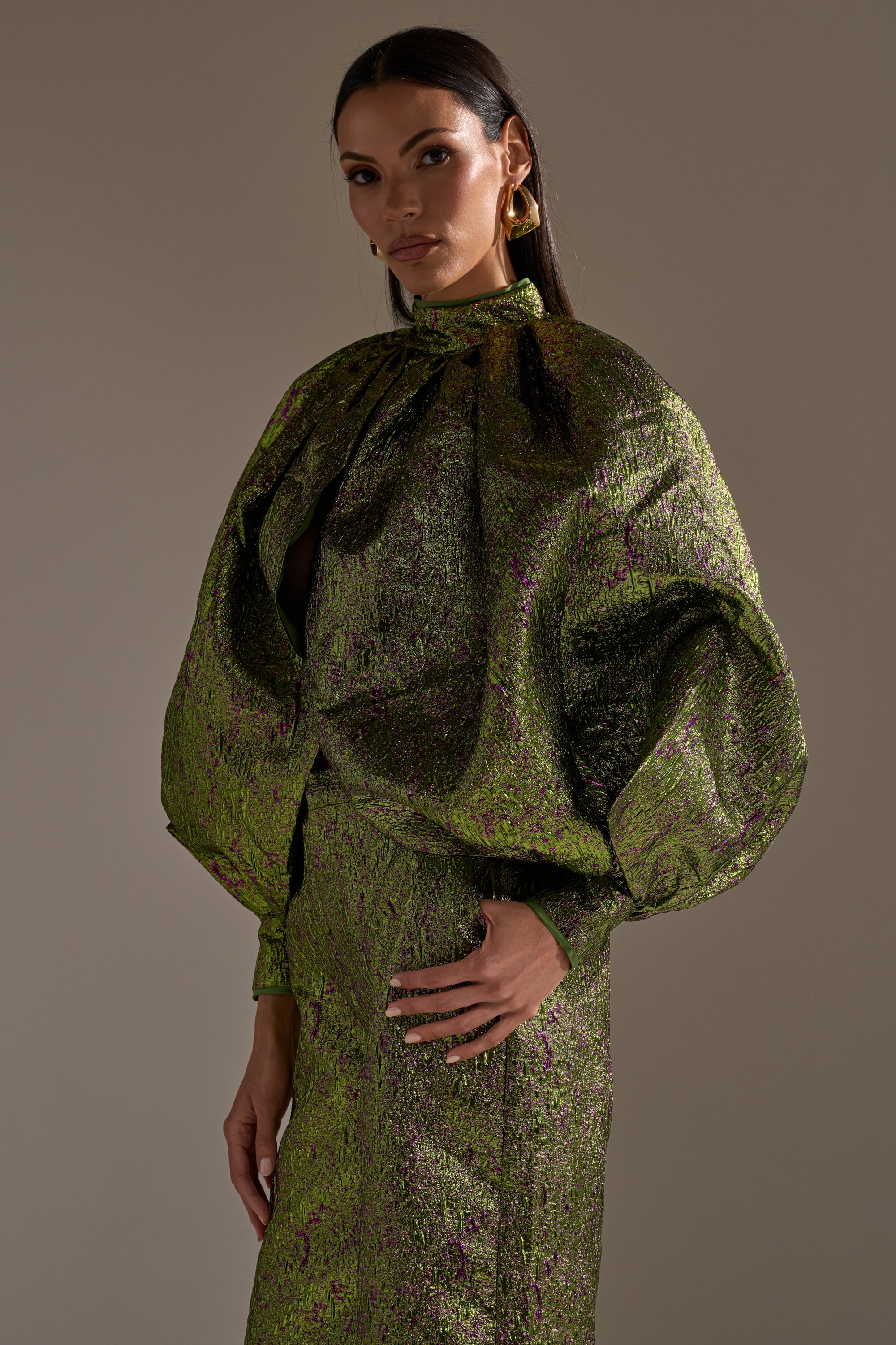 A woman models the LAYLA BROCADE PUFF SLEEVE MOCK NECK TOP in shiny green and purple, featuring voluminous puff sleeves and a cropped silhouette. She pairs it with gold earrings, long dark hair, and a confident look against a plain background.