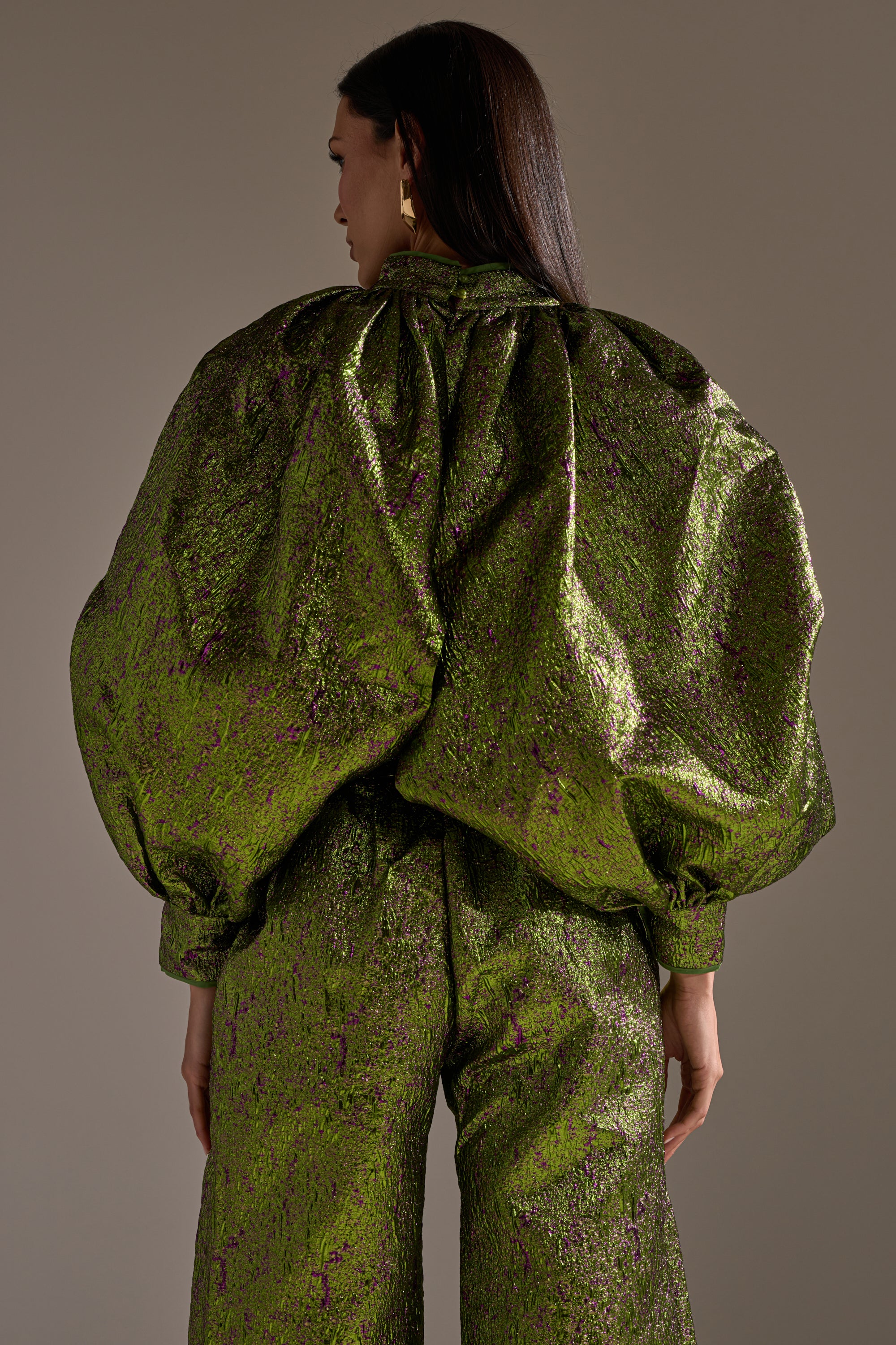 A person faces away, wearing the LAYLA BROCADE PUFF SLEEVE MOCK NECK TOP with voluminous sleeves and a shiny green finish. The cropped silhouette stands out against a plain background, emphasizing the bold, metallic statement piece.