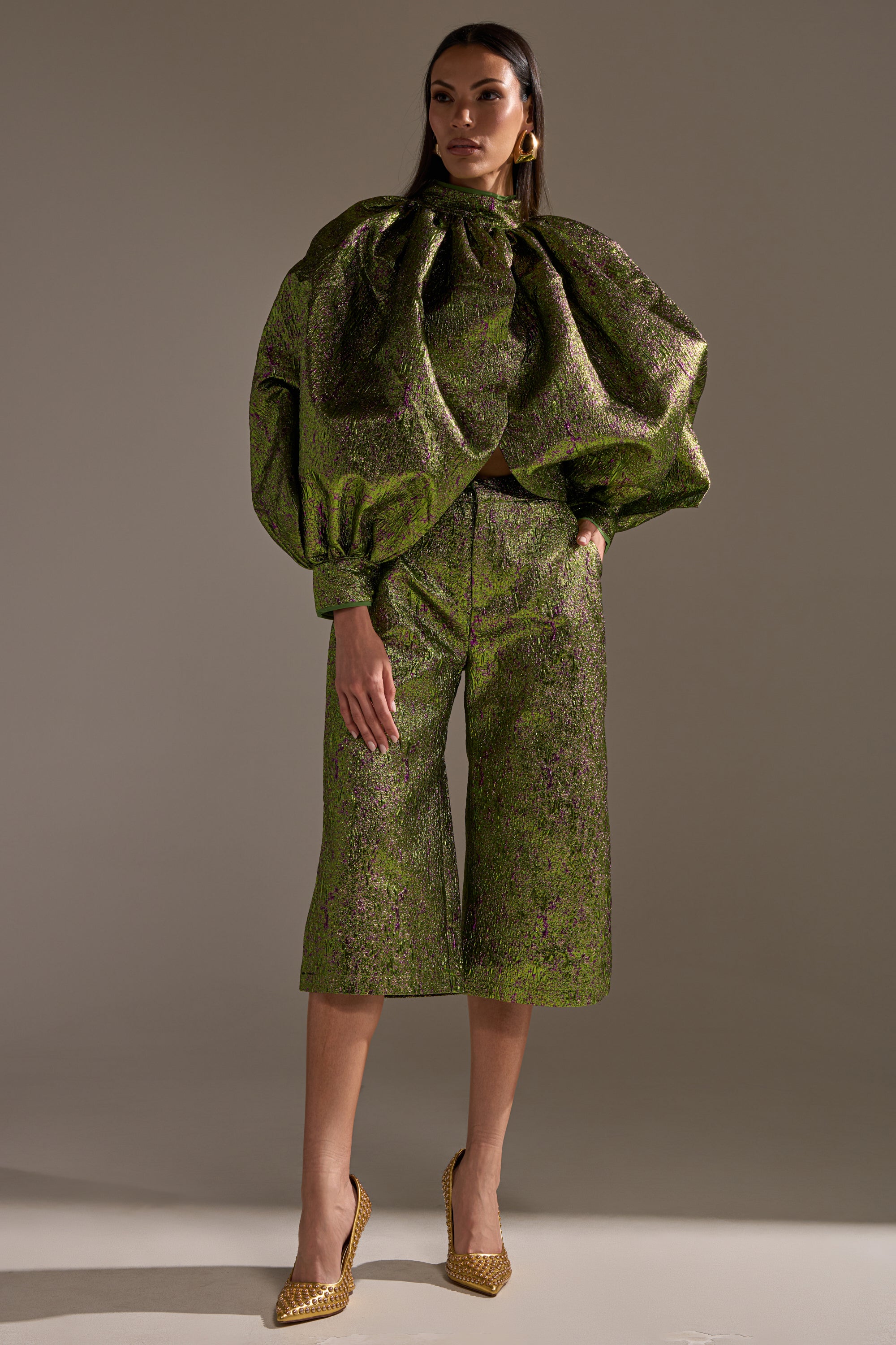 A woman models the LAYLA BROCADE PUFF SLEEVE MOCK NECK TOP in shiny green, paired with matching wide-leg cropped pants and gold pointed heels, standing out against a neutral background.