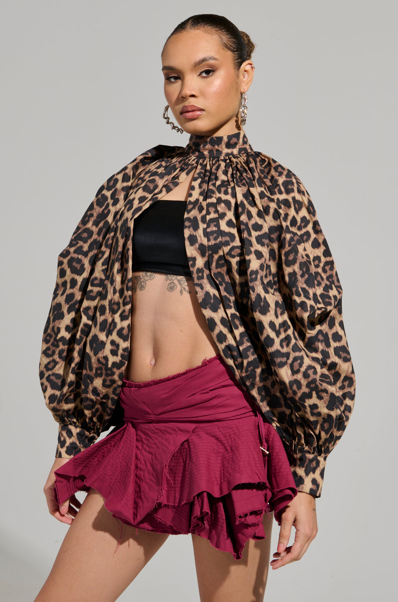A woman confidently showcases the DANGEROUS GIRL Leopard Long Sleeve Puff Blouse, paired with a maroon ruffled skirt. She stands against a neutral background, accessorized with large hoop earrings.