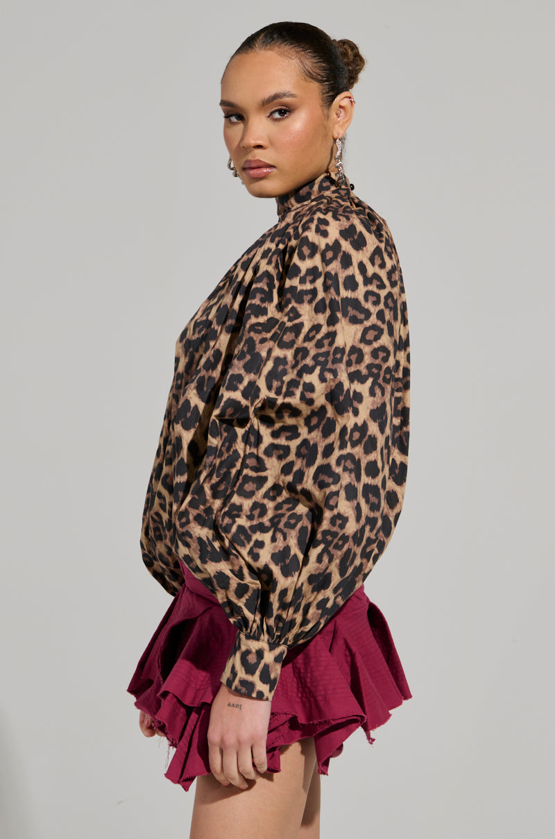 A woman poses against a neutral background, wearing the DANGEROUS GIRL LEOPARD LONG SLEEVE PUFF BLOUSE paired with a short, layered magenta skirt. Her hair is tied back, and she accessorizes with bold earrings. She gazes confidently towards the camera.