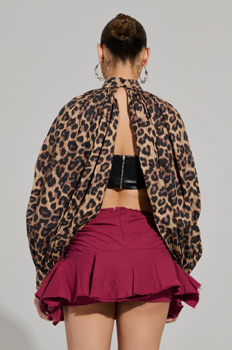 A person with their hair in a bun, adorned with hoop earrings, is wearing the DANGEROUS GIRL LEOPARD LONG SLEEVE PUFF BLOUSE featuring an open back and a cropped silhouette, paired with a burgundy pleated skirt. They are facing away from the camera against a gray background.