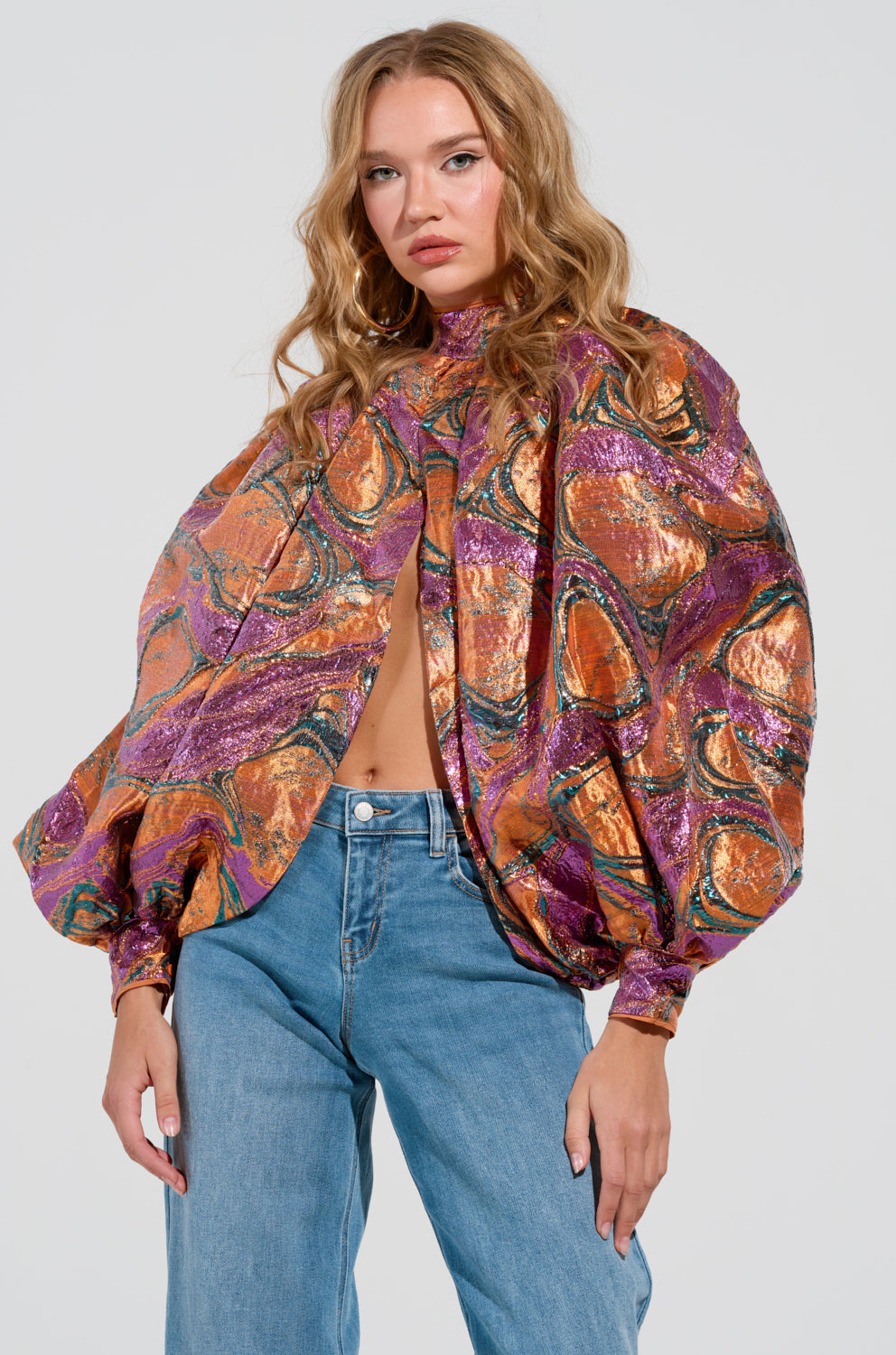 A person with long blonde hair is wearing the LUXURIOUS TENDENCIES BROCADE LONG SLEEVE PUFF BLOUSE, which features vibrant metallic patterns in shades of orange, purple, and green on textured fabric. The oversized blouse is paired with blue jeans against a plain white background.