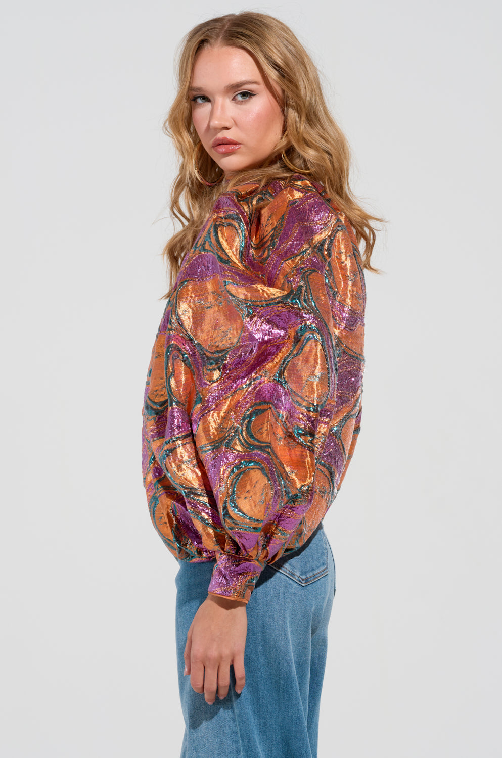 The woman with long wavy hair is wearing the LUXURIOUS TENDENCIES BROCADE LONG SLEEVE PUFF BLOUSE, which features a colorful abstract pattern, along with blue jeans. She stands in profile against a plain background, embodying an air of elegance with her neutral expression.
