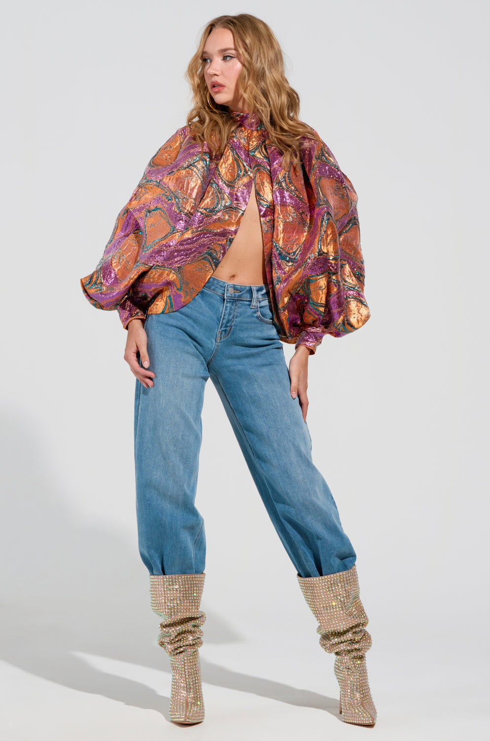 A person with long, wavy hair stands confidently in the Luxurious Tendencies Brocade Long Sleeve Puff Blouse, paired with high-waisted blue jeans and sparkling knee-high boots, exuding elegance against a plain white background.