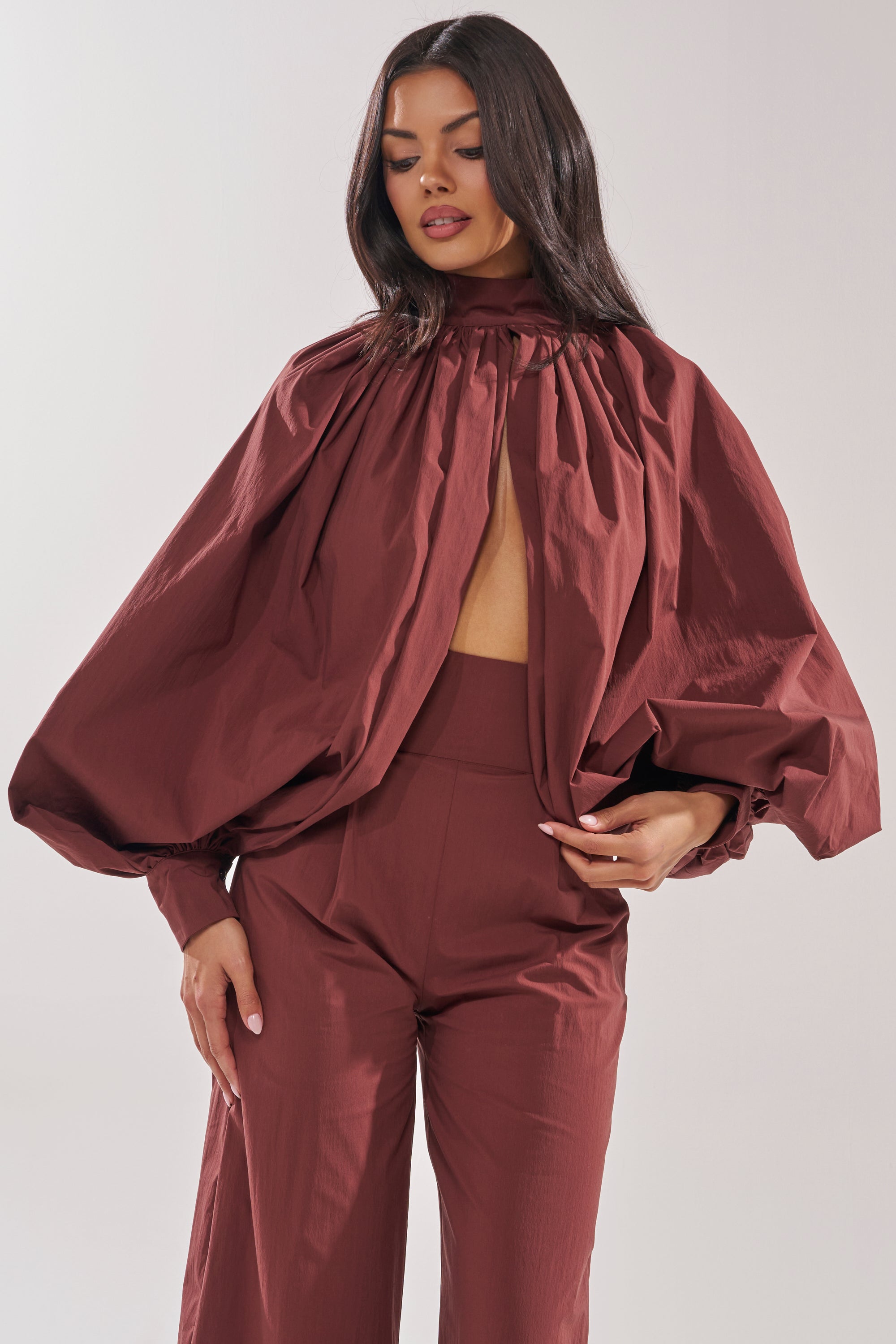 A woman poses confidently, wearing the GOLD COAST BALLOON SLEEVE BLOUSE IN BROWN with a high neck and front cutout, paired with matching high-waisted pants. She has long dark hair and rests one hand on her waist. 0