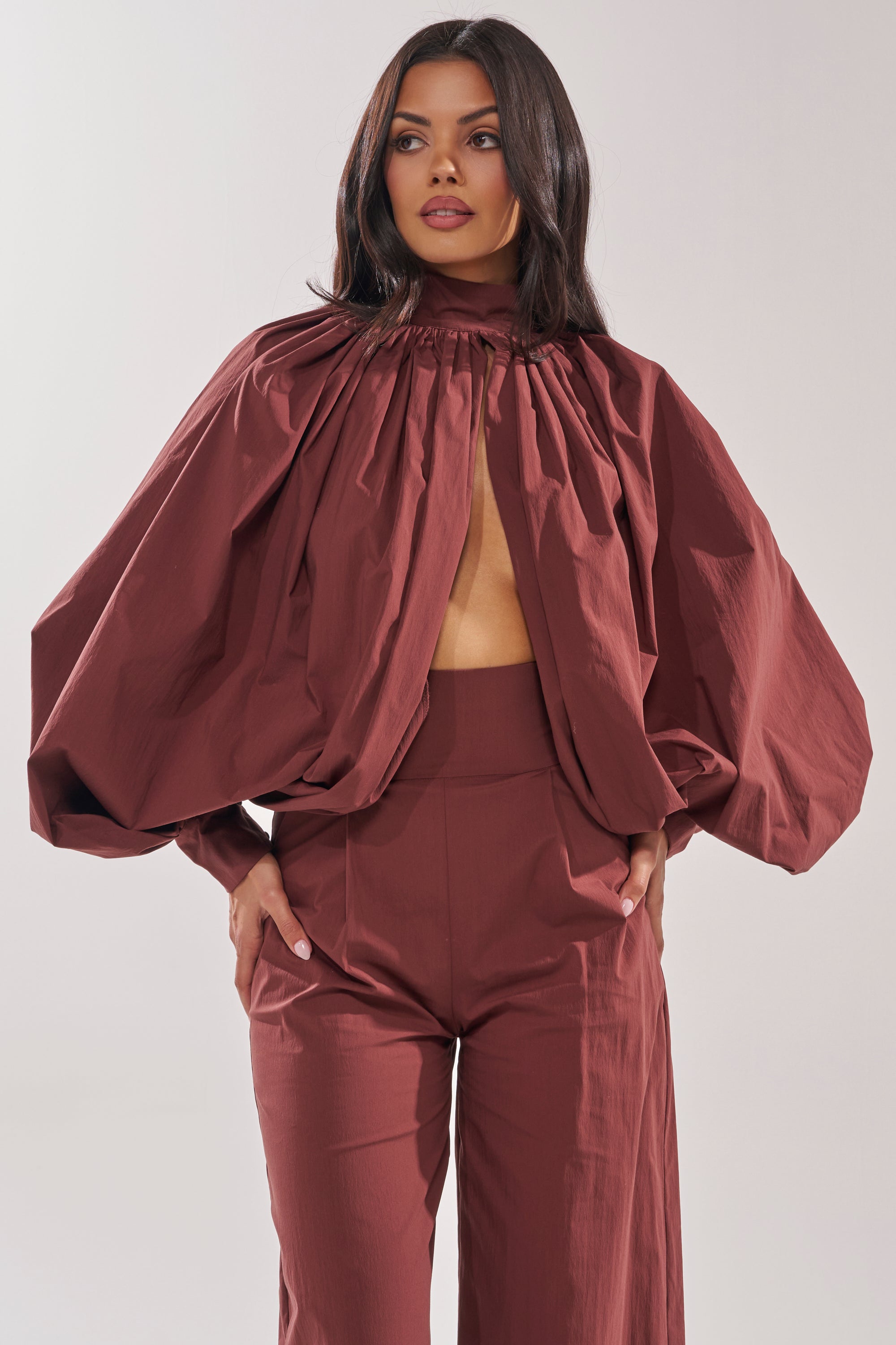 A woman with long, dark hair and a neutral expression wears the GOLD COAST BALLOON SLEEVE BLOUSE IN BROWN featuring voluminous balloon sleeves, a gathered neckline, and an open front that reveals her midriff, paired with wide-leg high-waisted pants. 1