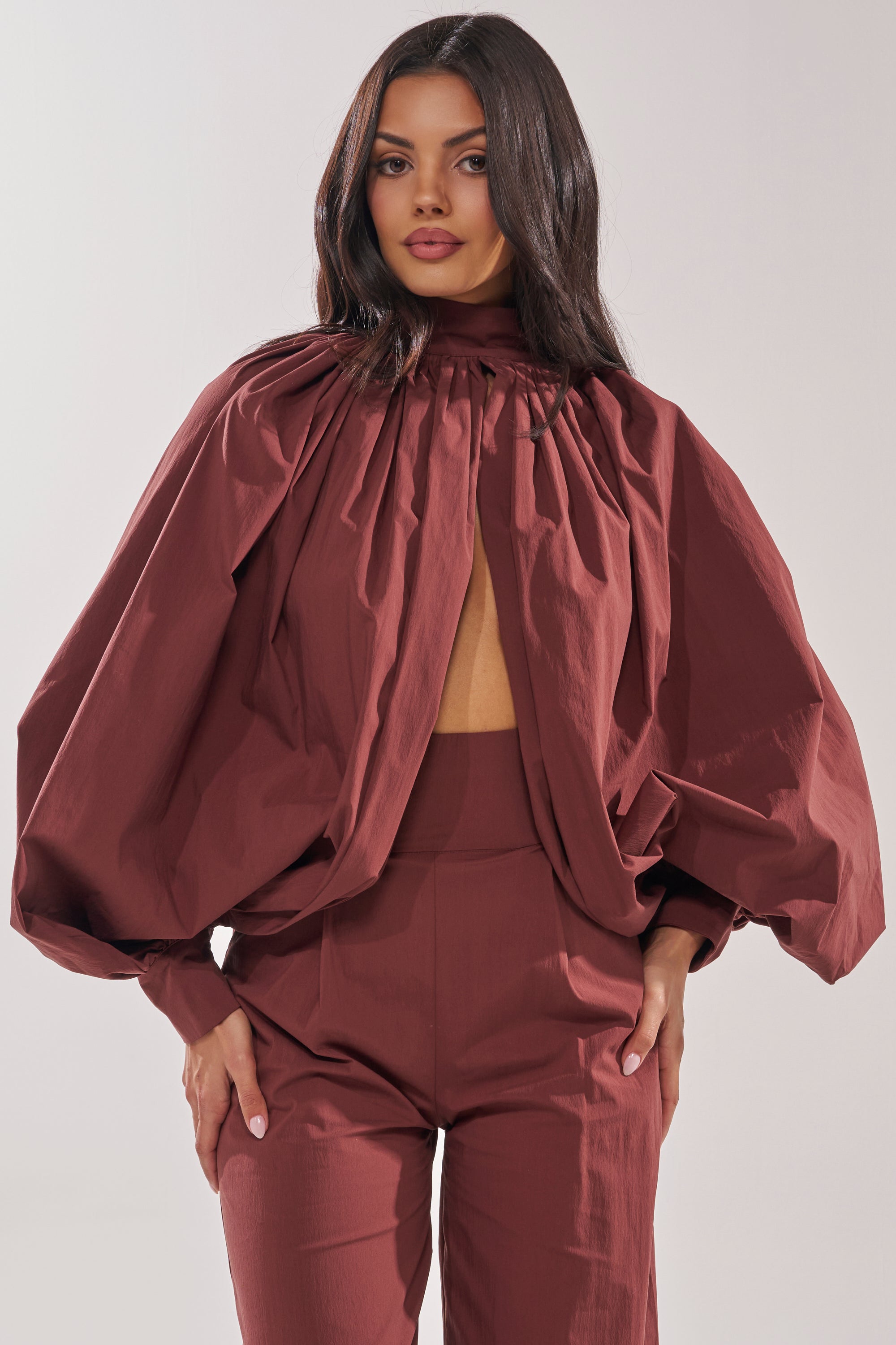 A woman wears the GOLD COAST BALLOON SLEEVE BLOUSE IN BROWN, a high-neck top with voluminous balloon sleeves and open front, paired with matching high-waisted pants, embodying modern minimalism against a plain light background. 2