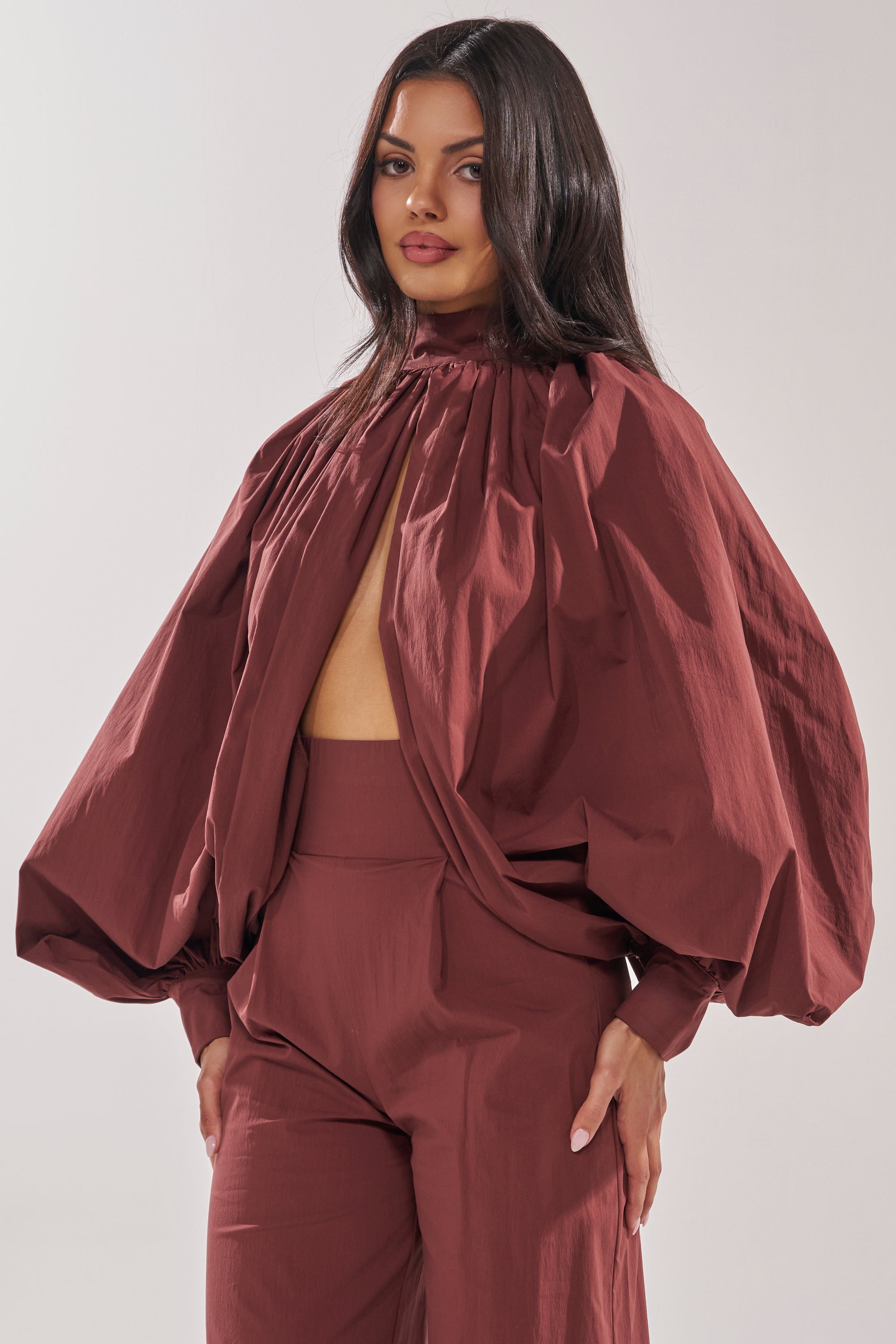 A woman stands confidently against a plain background in the GOLD COAST BALLOON SLEEVE BLOUSE IN BROWN, featuring a stylish open back and voluminous sleeves, paired with high-waisted pants. 3