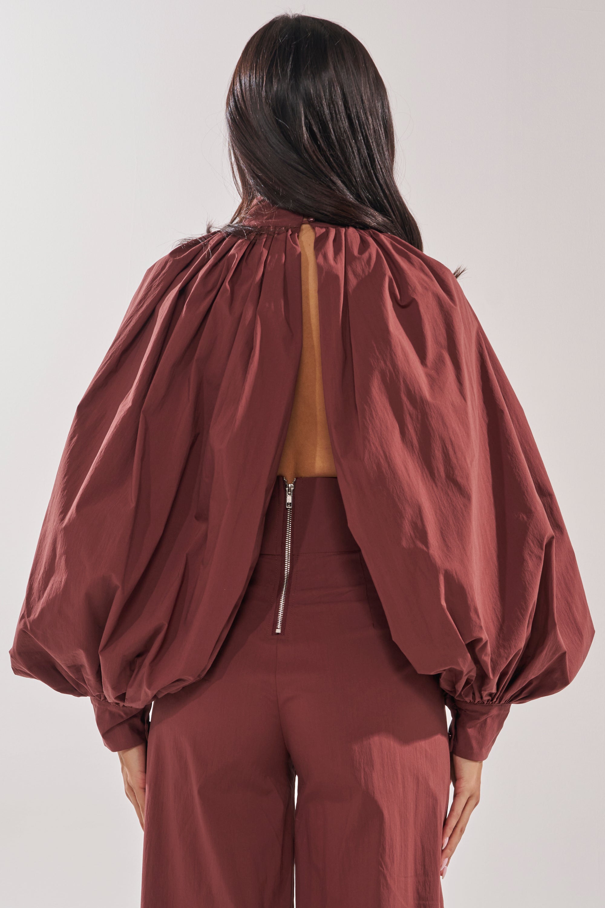 A person with long dark hair is seen from the back wearing the GOLD COAST BALLOON SLEEVE BLOUSE IN BROWN, featuring a loose fit and open-back design for added intrigue, paired with pants that have a visible back zipper for chic style. 4