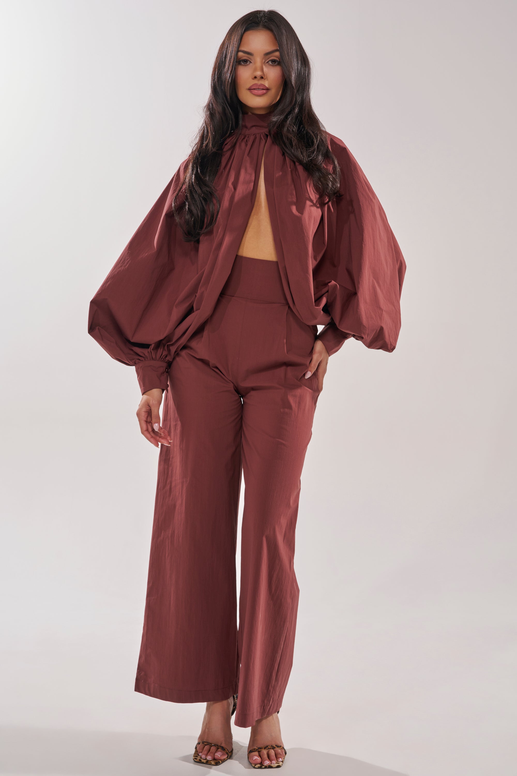 A woman radiates confidence in the GOLD COAST BALLOON SLEEVE BLOUSE IN BROWN, styled with wide-leg pants and open-toe heels, standing against a plain backdrop for a modern minimalist look. 7