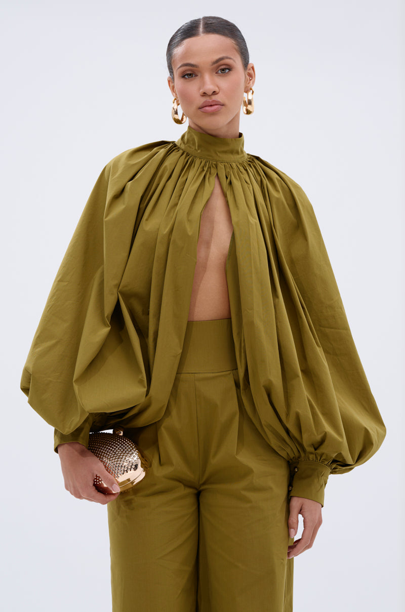 A woman models the GOLD COAST BALLOON SLEEVE BLOUSE in olive green with matching trousers, styled with a gold clutch, slicked-back hair, and large gold hoop earrings against a plain backdrop.