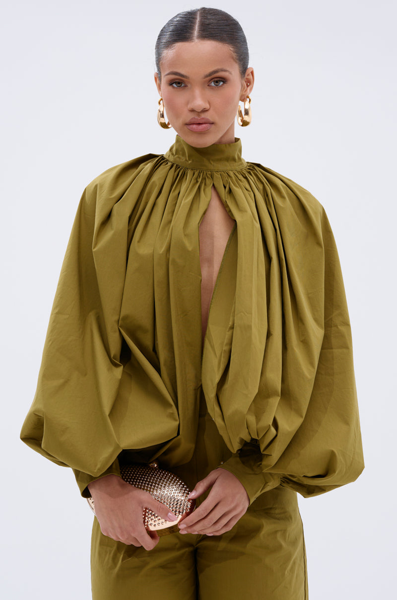 A woman with slicked-back hair wears the GOLD COAST BALLOON SLEEVE BLOUSE in olive green, featuring a high neck and voluminous sleeves. She accessorizes with large gold hoops and a textured gold clutch against a plain backdrop.