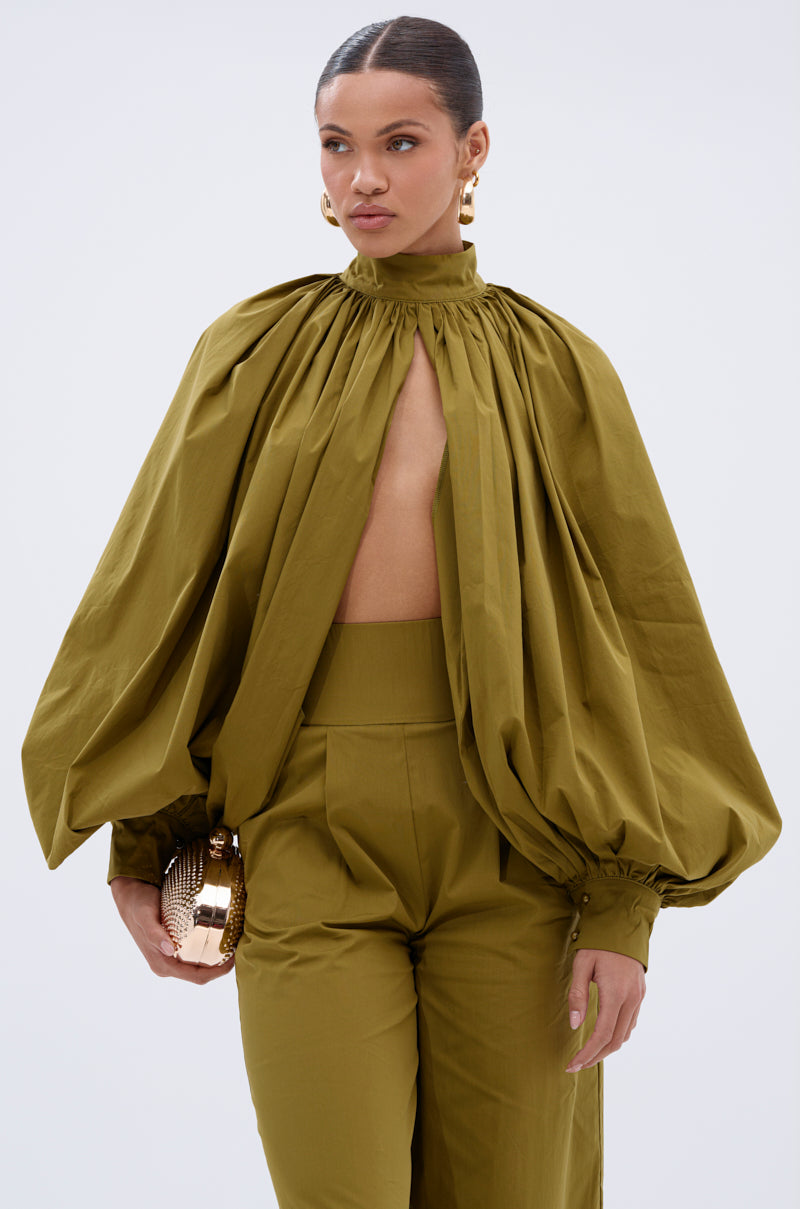 A woman wears the GOLD COAST BALLOON SLEEVE BLOUSE in olive green, featuring billowy sleeves and an open front. She pairs it with high-waisted pants, gold earrings, a gold clutch, and has her hair tied back.