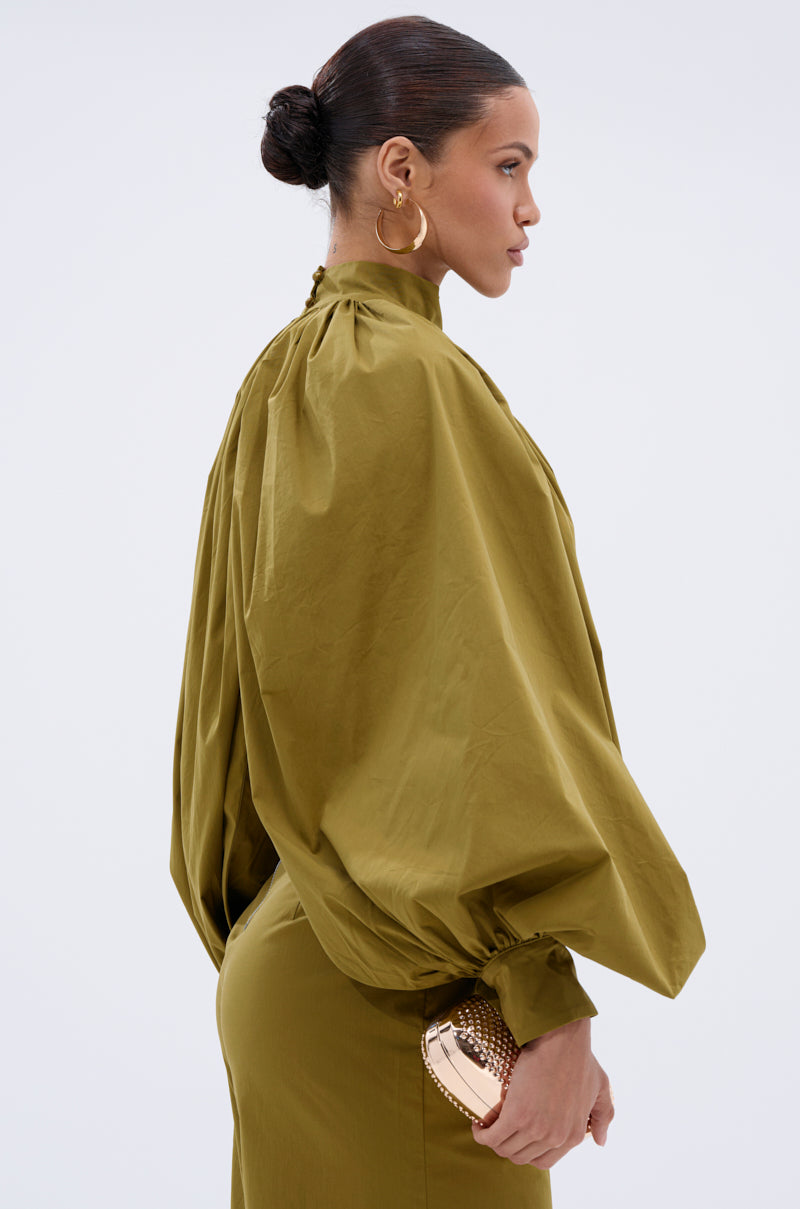 A woman stands in profile against a light background, wearing the GOLD COAST BALLOON SLEEVE BLOUSE in olive green with a high neck. She accessorizes with large hoop earrings, a gold clutch, and has her hair pulled back in a sleek bun.