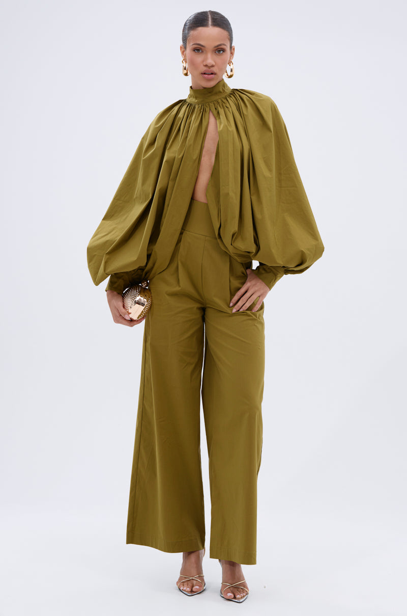 A woman wears the GOLD COAST BALLOON SLEEVE BLOUSE—an olive green, high-neck top with voluminous sleeves—paired with matching wide-leg pants, gold clutch, heels, and statement earrings against a plain light background.